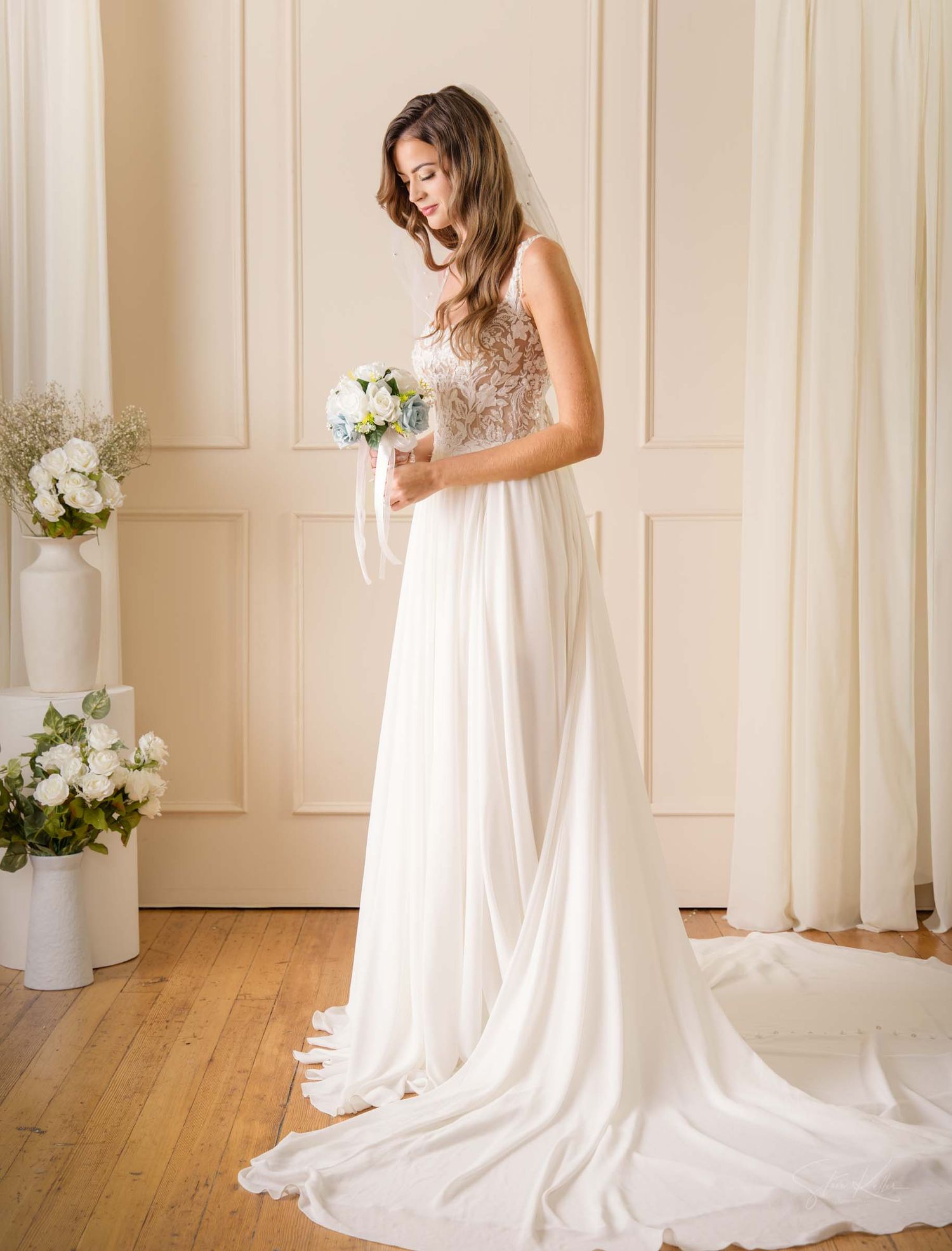 A flowing white wedding dress with a long train displayed in a bright studio setting with hardwood floors and cream walls.