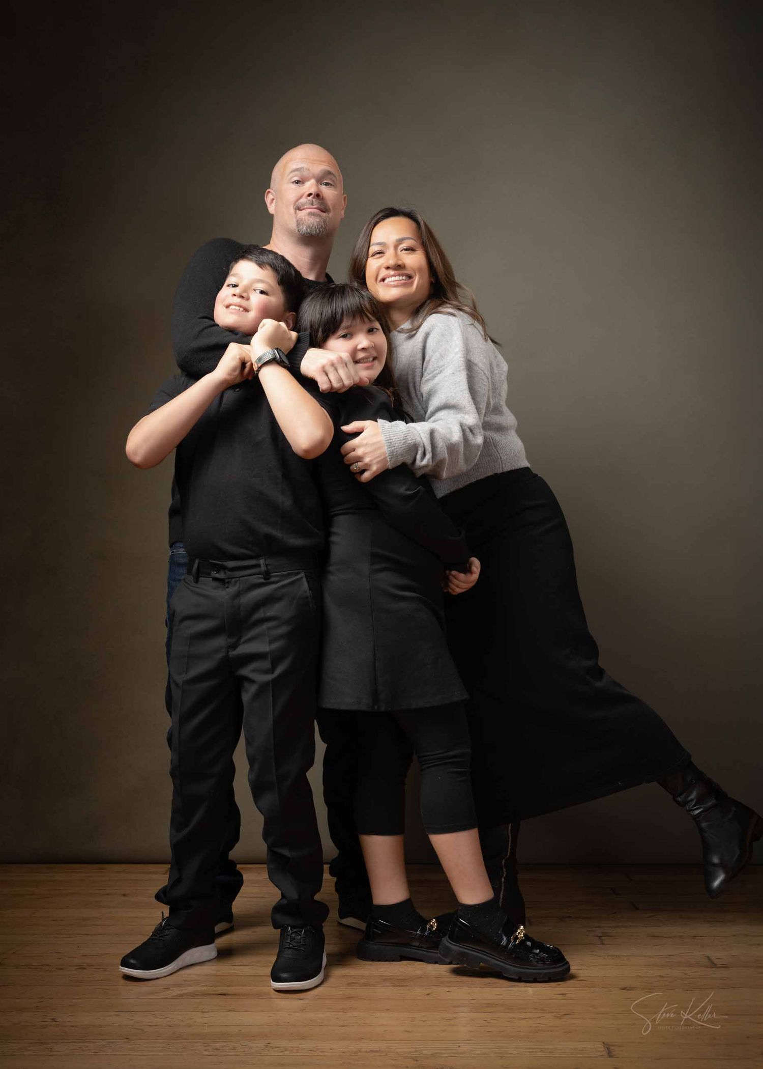 A family of four poses together against a dark backdrop in coordinated black and gray outfits on a wooden floor.
