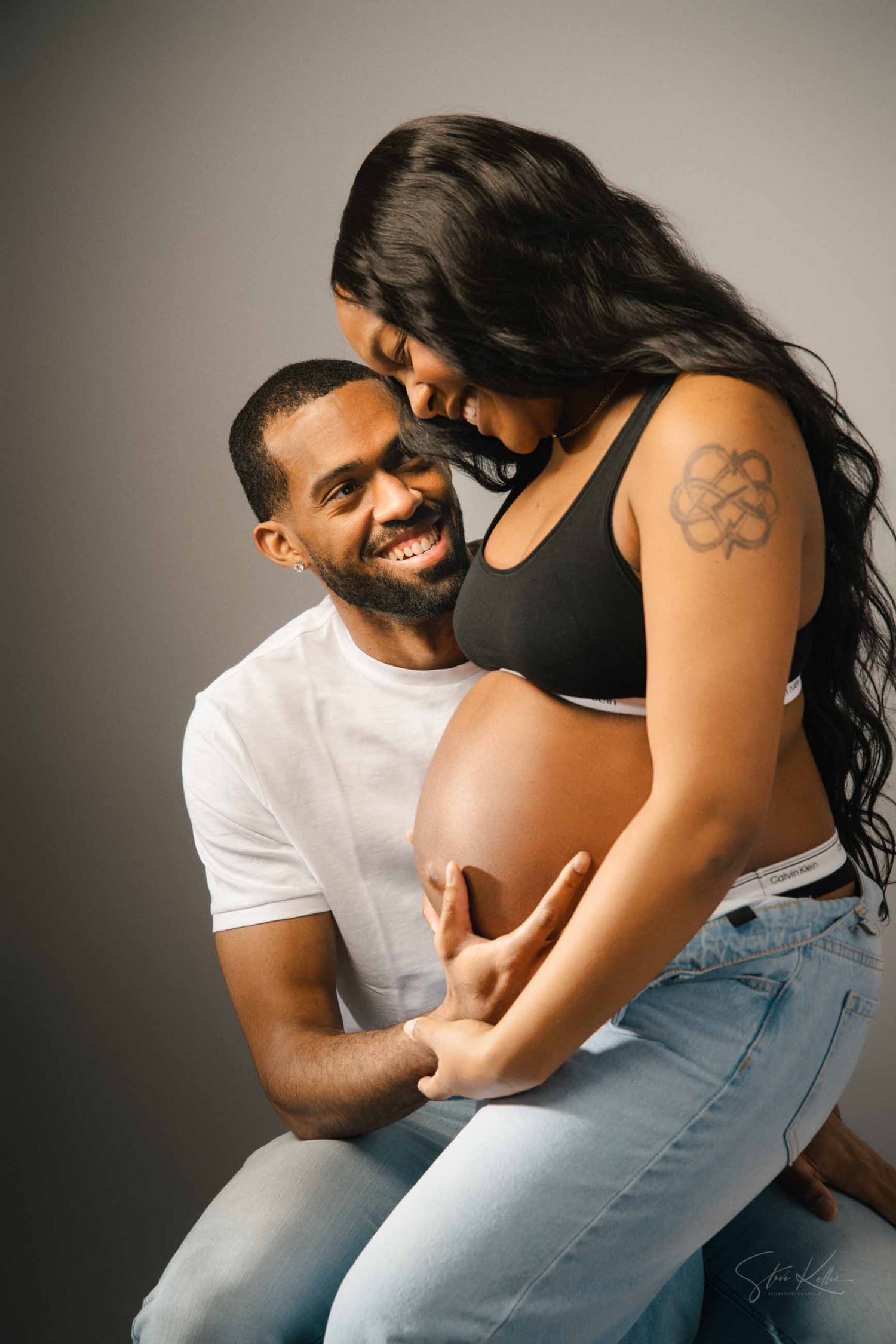 A couple embraces and smiles during a pregnancy photoshoot in a studio with neutral background.