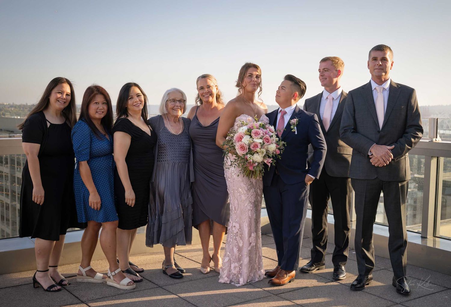Wedding party group photo on rooftop at sunset, bride holding pink bouquet with family and friends.