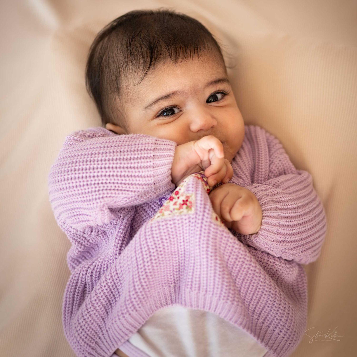 Adorable baby in a lavender knit sweater chewing on sleeve, looking up with big brown eyes on a soft beige background.