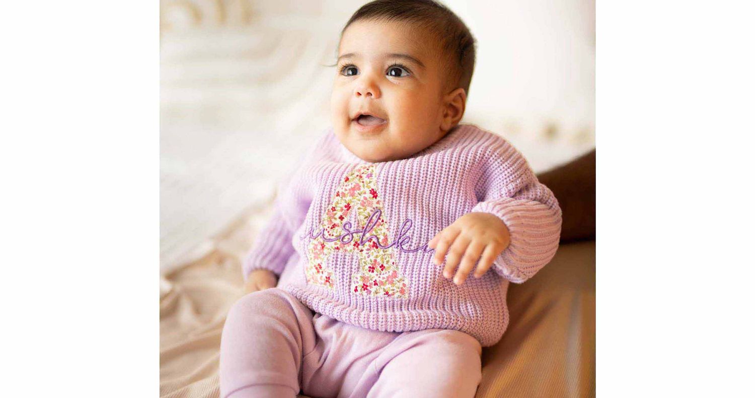 Cheerful baby in a lilac knit sweater with floral embroidery and matching pink leggings sits on a soft beige blanket.