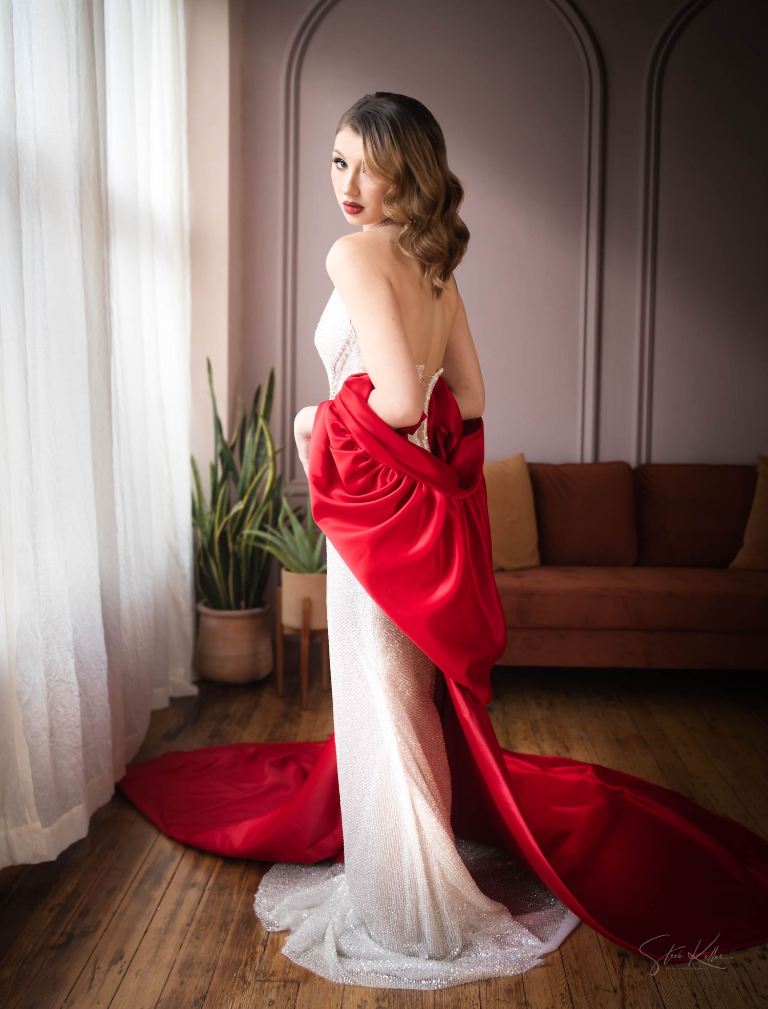A flowing red dress contrasts elegantly with a white gown against soft gray walls and wood flooring in a romantic indoor setting.