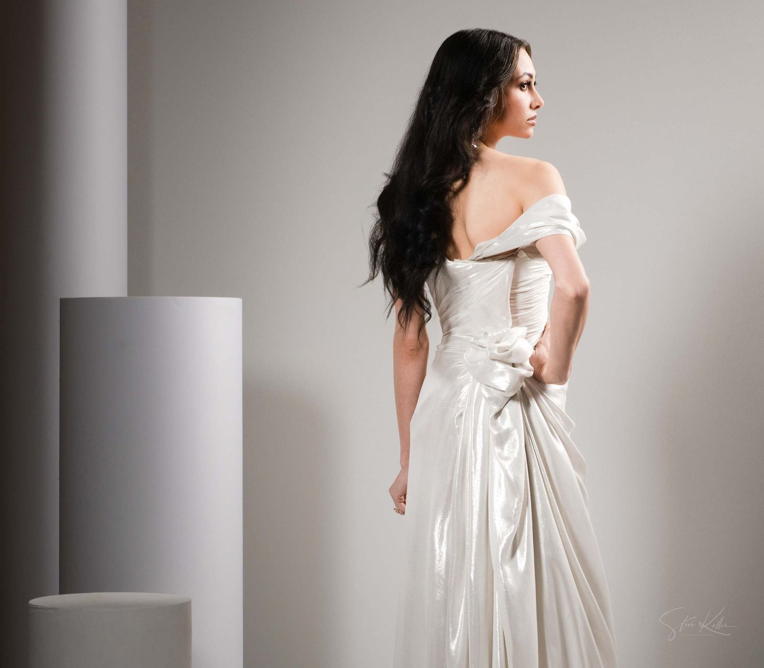 A woman in a white off-shoulder dress poses gracefully against a minimalist gray studio backdrop.