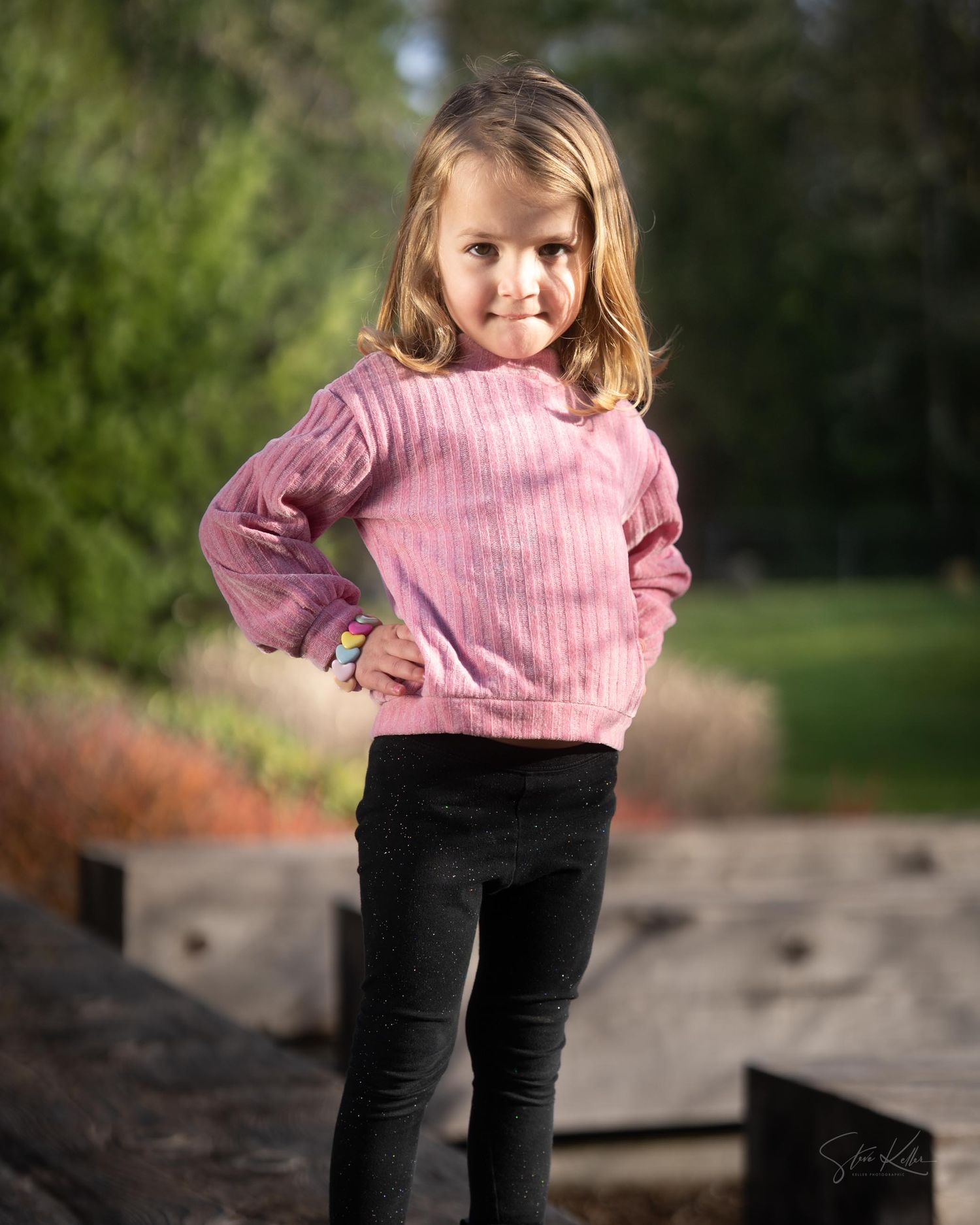 A child in a pink sweater and black pants poses playfully outdoors in an autumn setting.