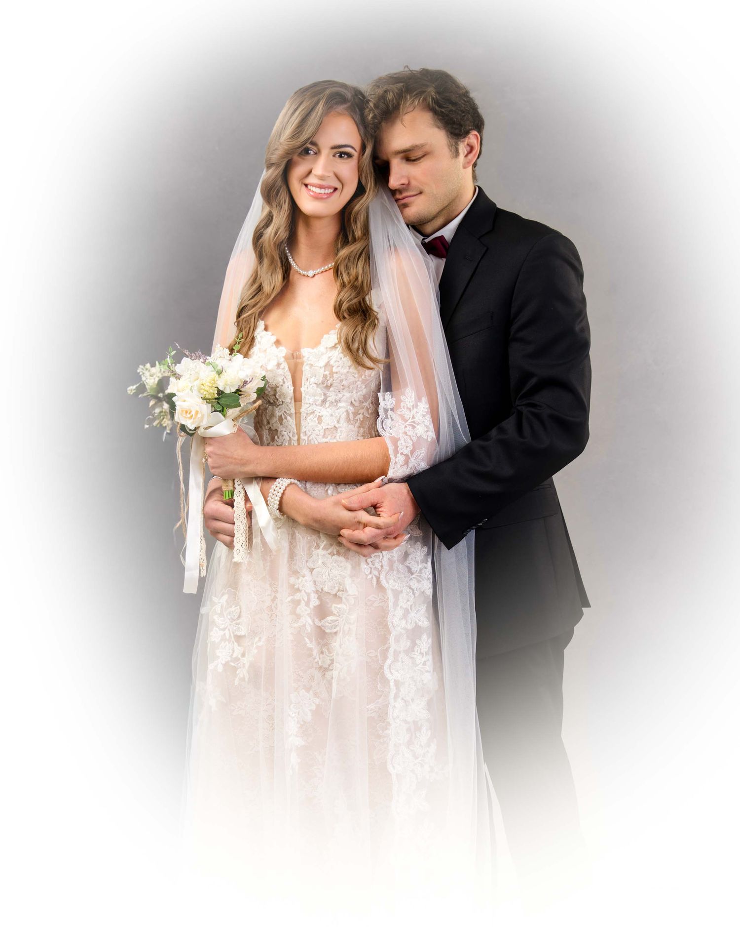 A romantic wedding couple embraces in soft lighting while wearing elegant white dress and black suit.
