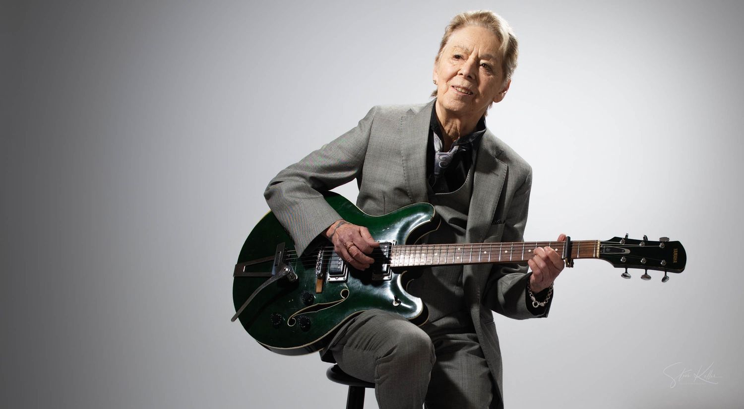 A person wearing a gray suit poses with a green electric guitar against a white background.