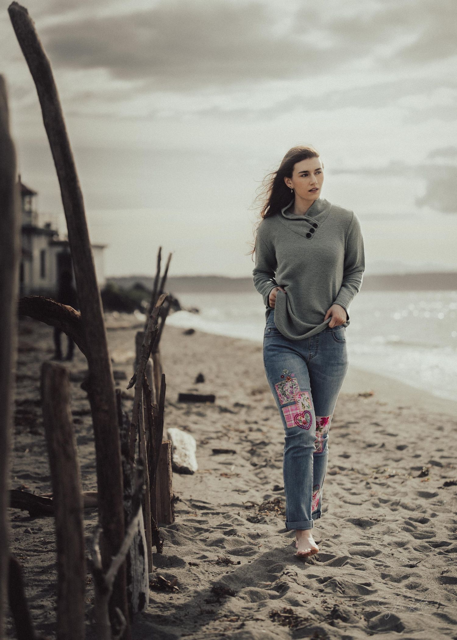A person in a gray sweater and patched jeans walking along a moody beach with driftwood on an overcast day.