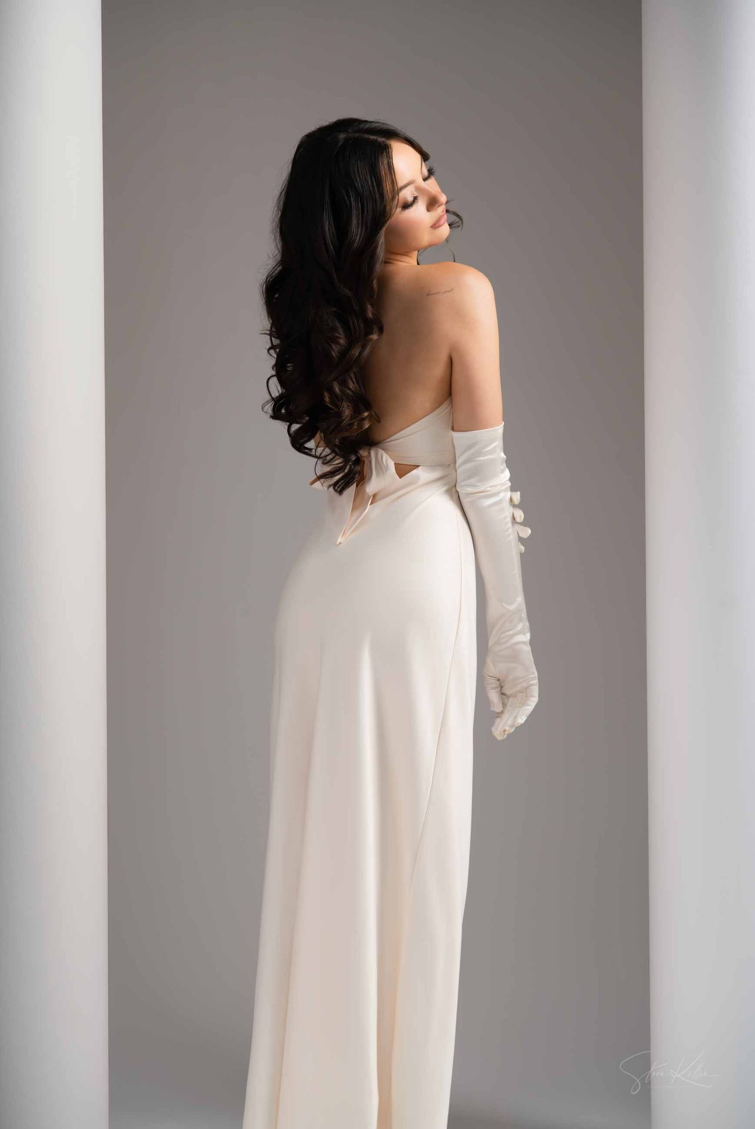 A woman in a flowing white dress and long gloves poses elegantly against a neutral grey studio backdrop.