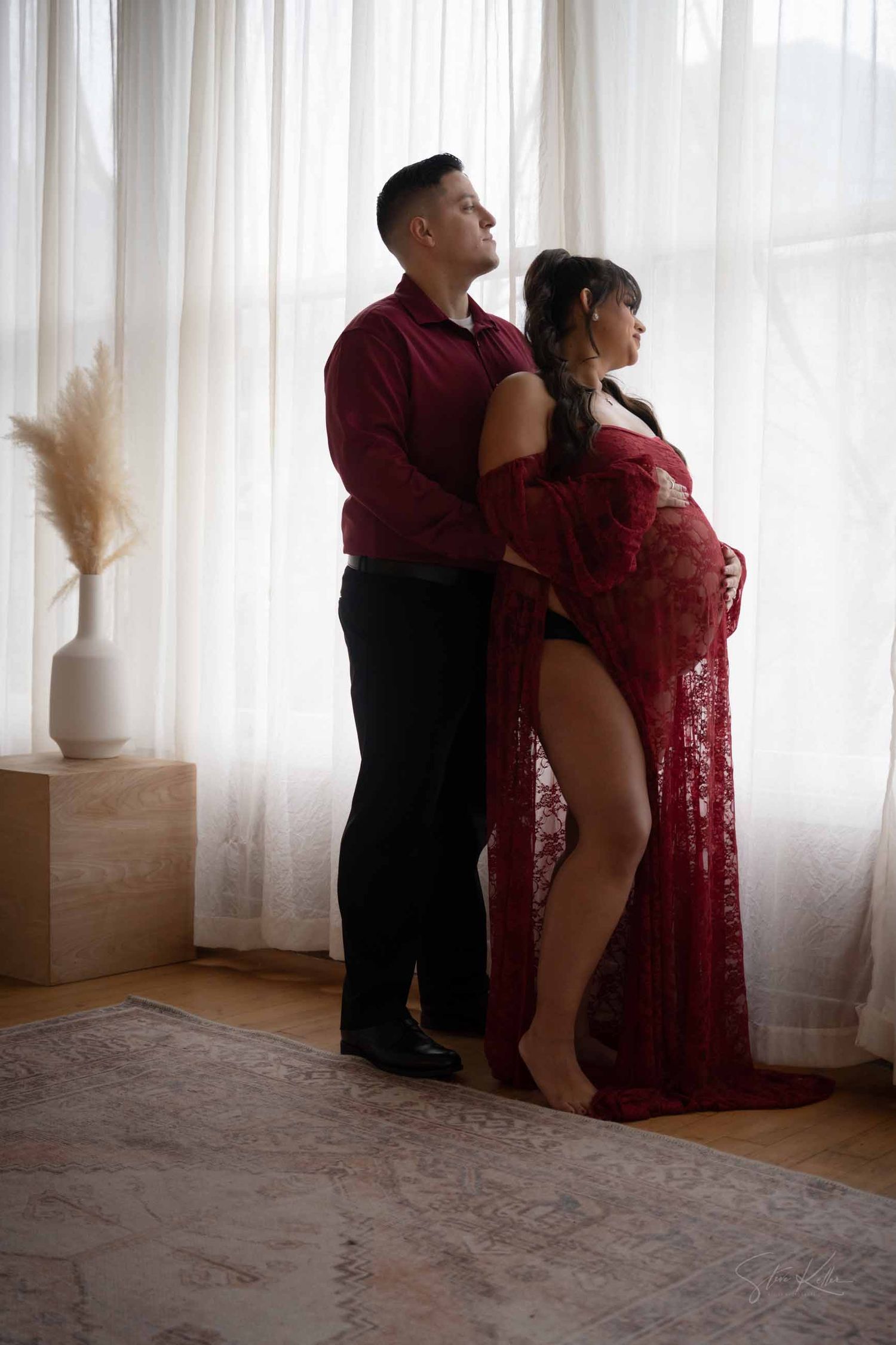 A couple in matching burgundy outfits poses together by sheer curtains in an intimate maternity photoshoot.