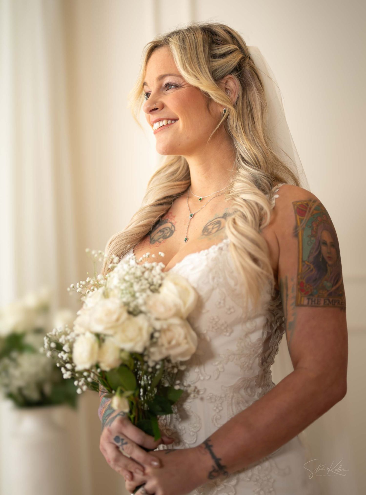 A bride with blonde hair and tattoos holds a white rose and baby's breath bouquet while smiling.