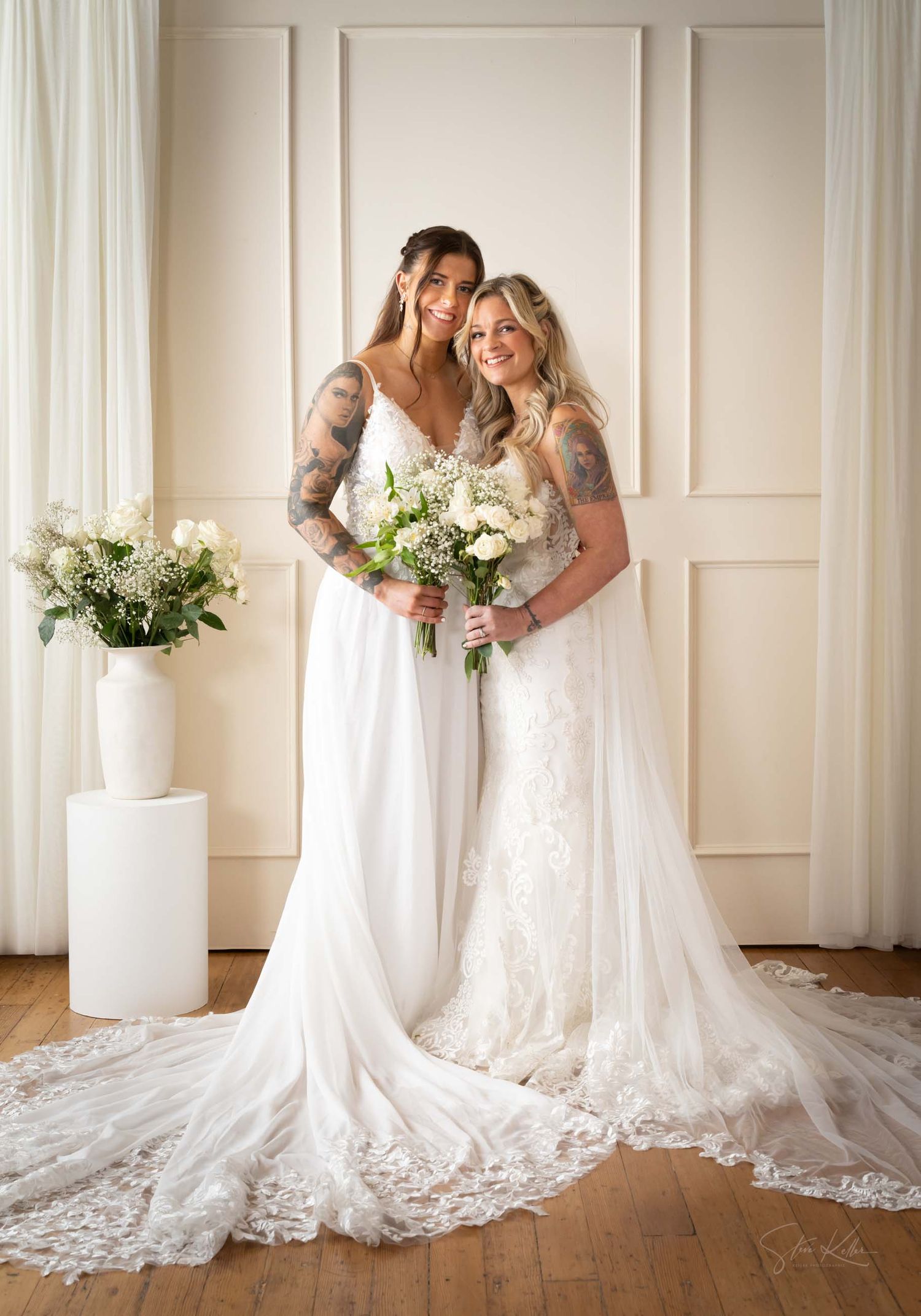 Two people in elegant white wedding dresses with long lace trains pose together holding white floral bouquets.