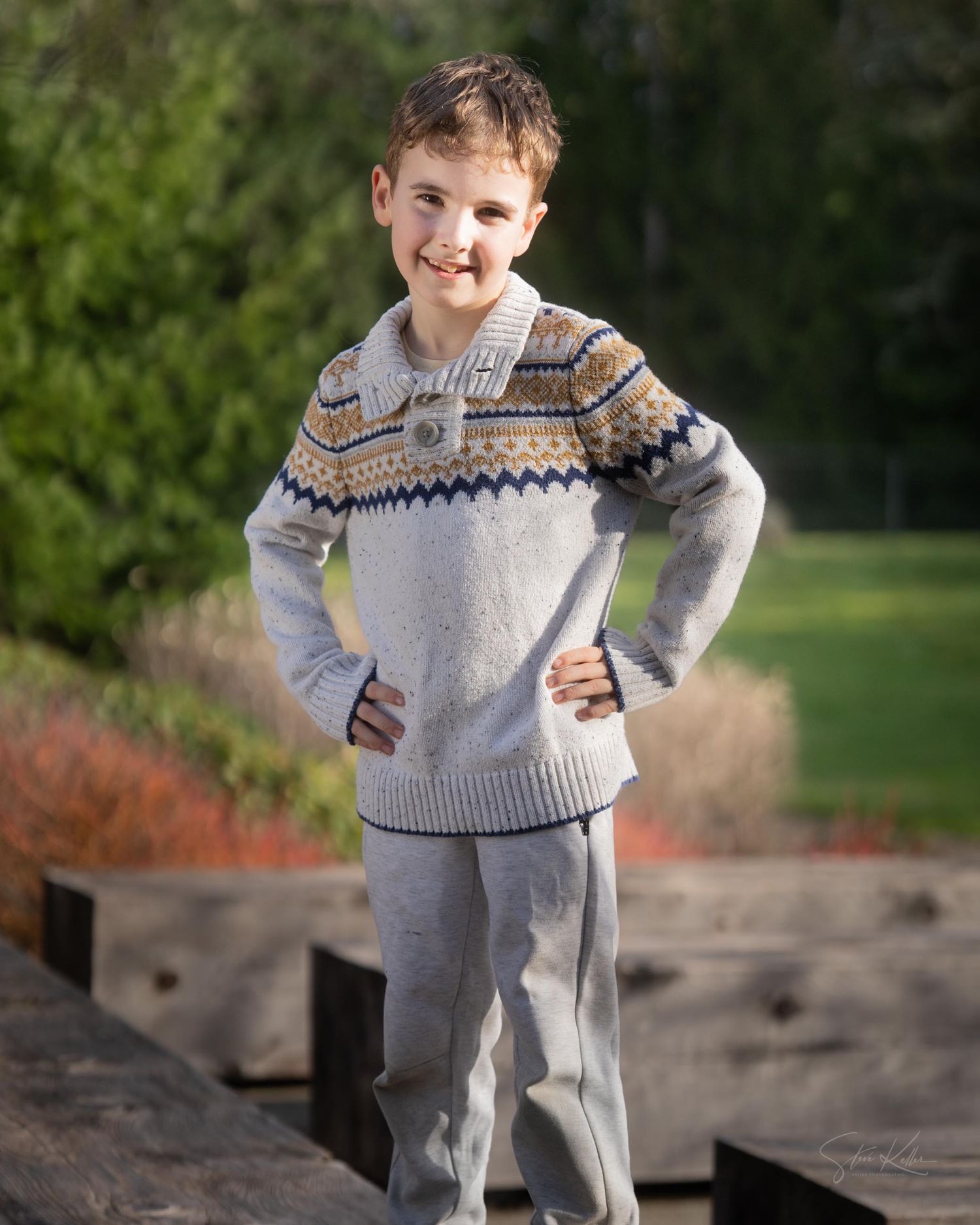 A young person in a patterned winter sweater and light colored pants stands confidently on wooden decking outdoors.