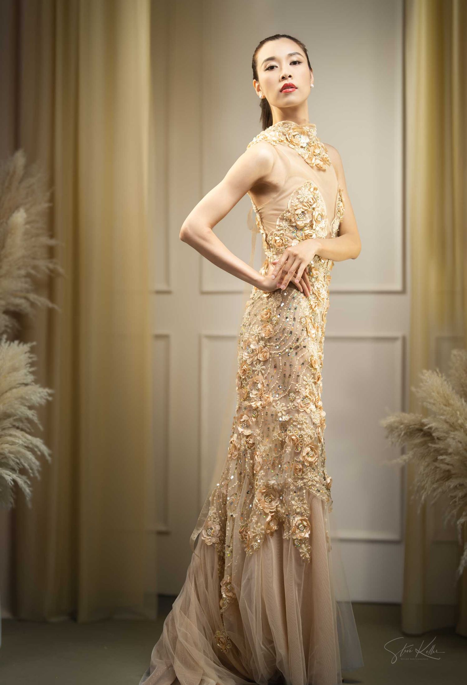 A model poses in an elegant beige evening gown with delicate lace details and flowing tulle skirt in a studio setting.