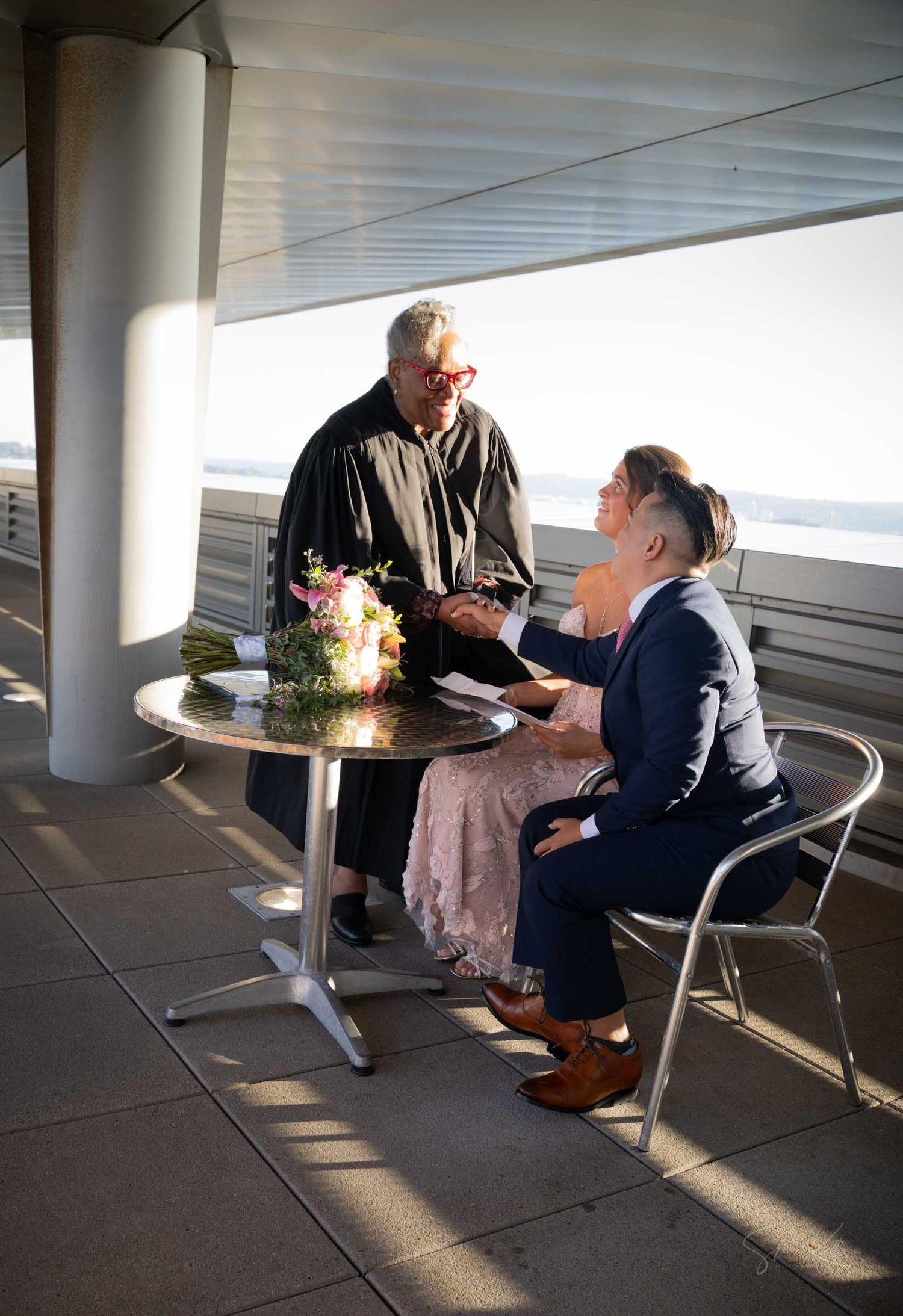 Wedding officiant in black robe reviews documents with groom at outdoor waterfront table before ceremony.