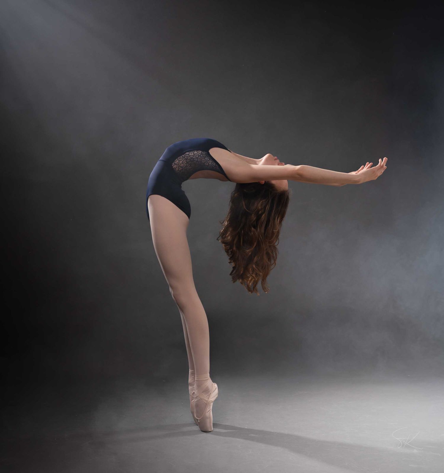 Ballet dancer en pointe in blue leotard performs a dramatic back arch with arms extended in studio.