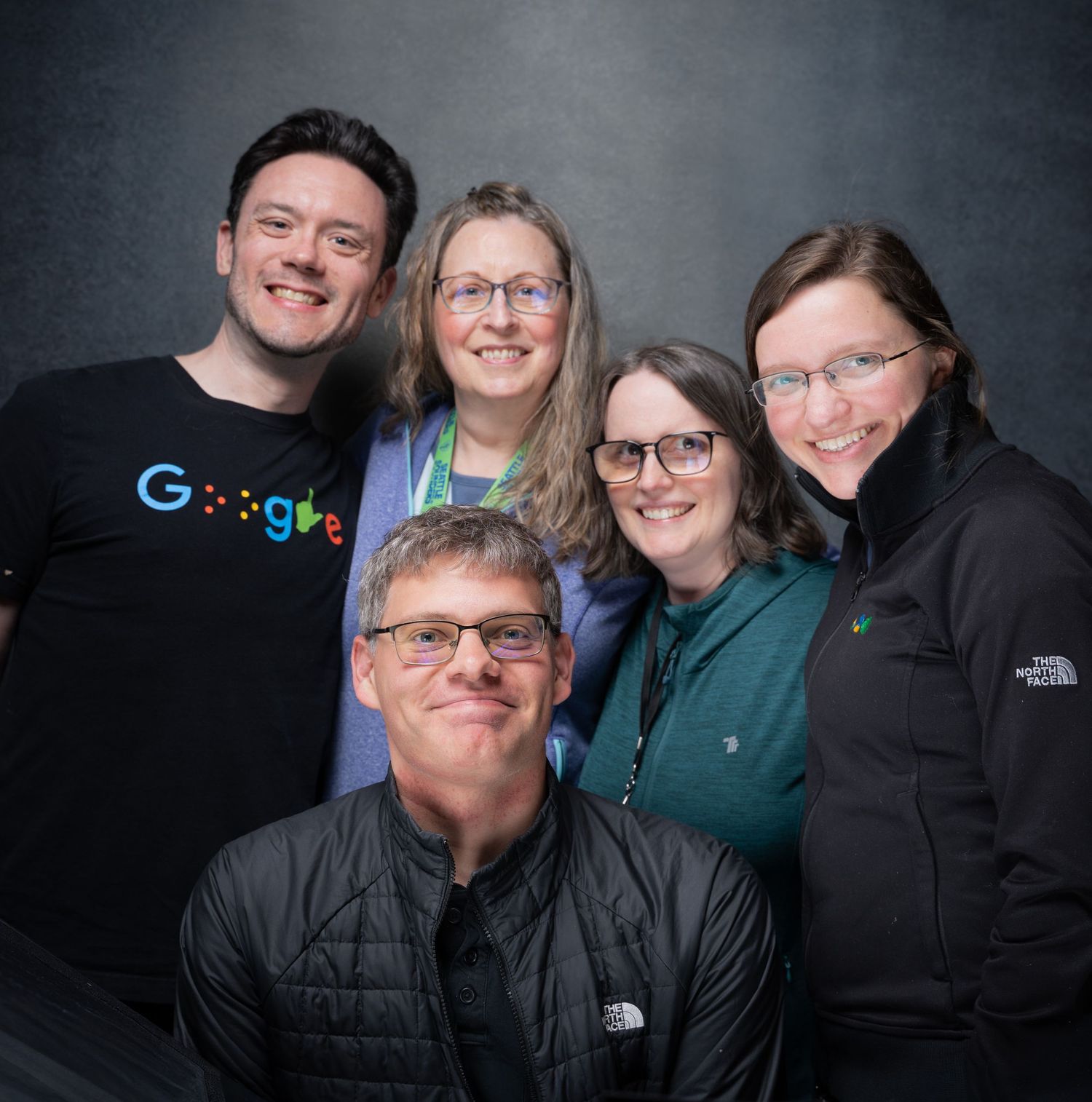 Five smiling adults pose together for a group photo, one wearing a Google t-shirt, in a casual portrait setting.