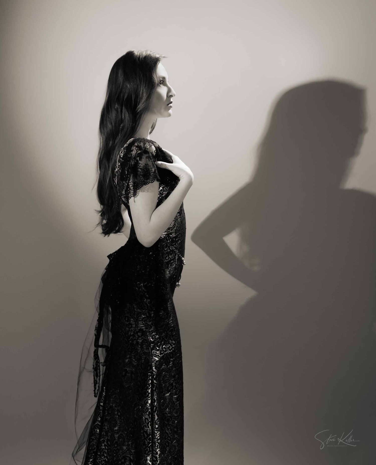 Silhouetted figure in black lace dress poses against white wall creating dramatic shadow play.