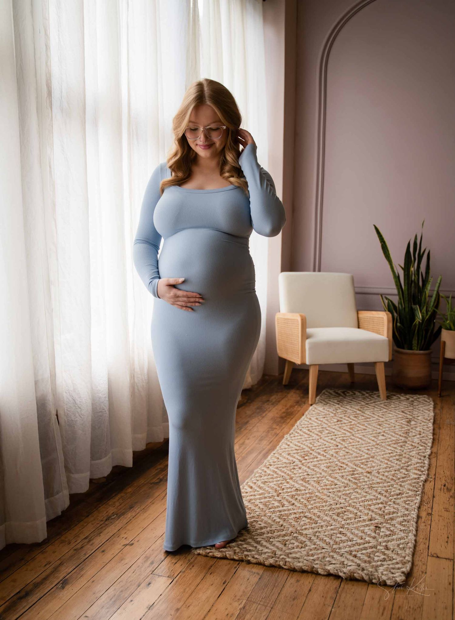 A pregnant woman in a fitted blue long sleeve dress poses by a window next to a modern accent chair and houseplant.