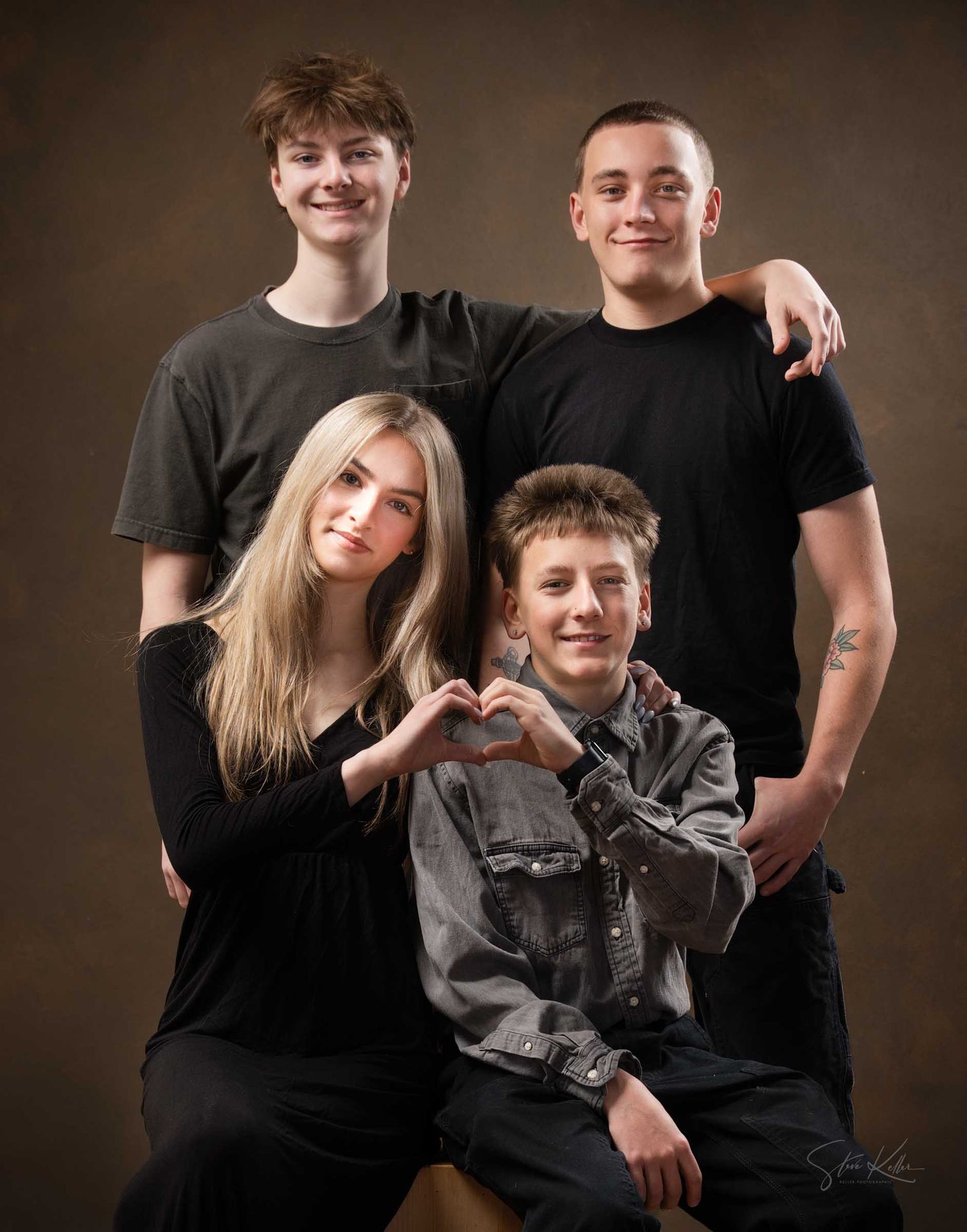 Four teenagers pose for a professional studio portrait, with two boys standing behind a girl and boy making a heart shape.