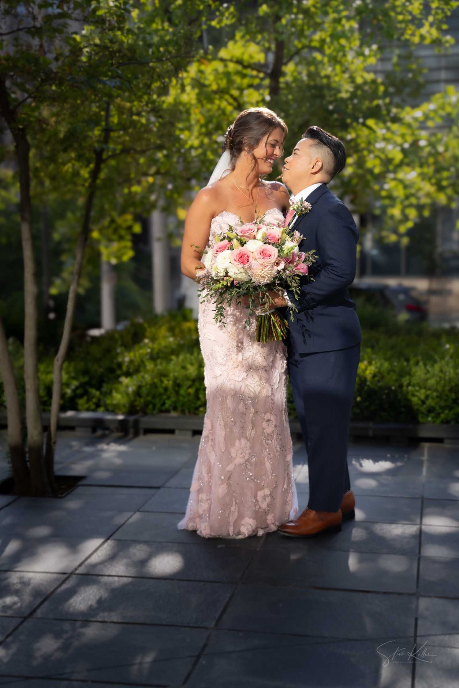 Bride in lace gown holding pink bouquet shares romantic moment with groom in navy suit outdoors.