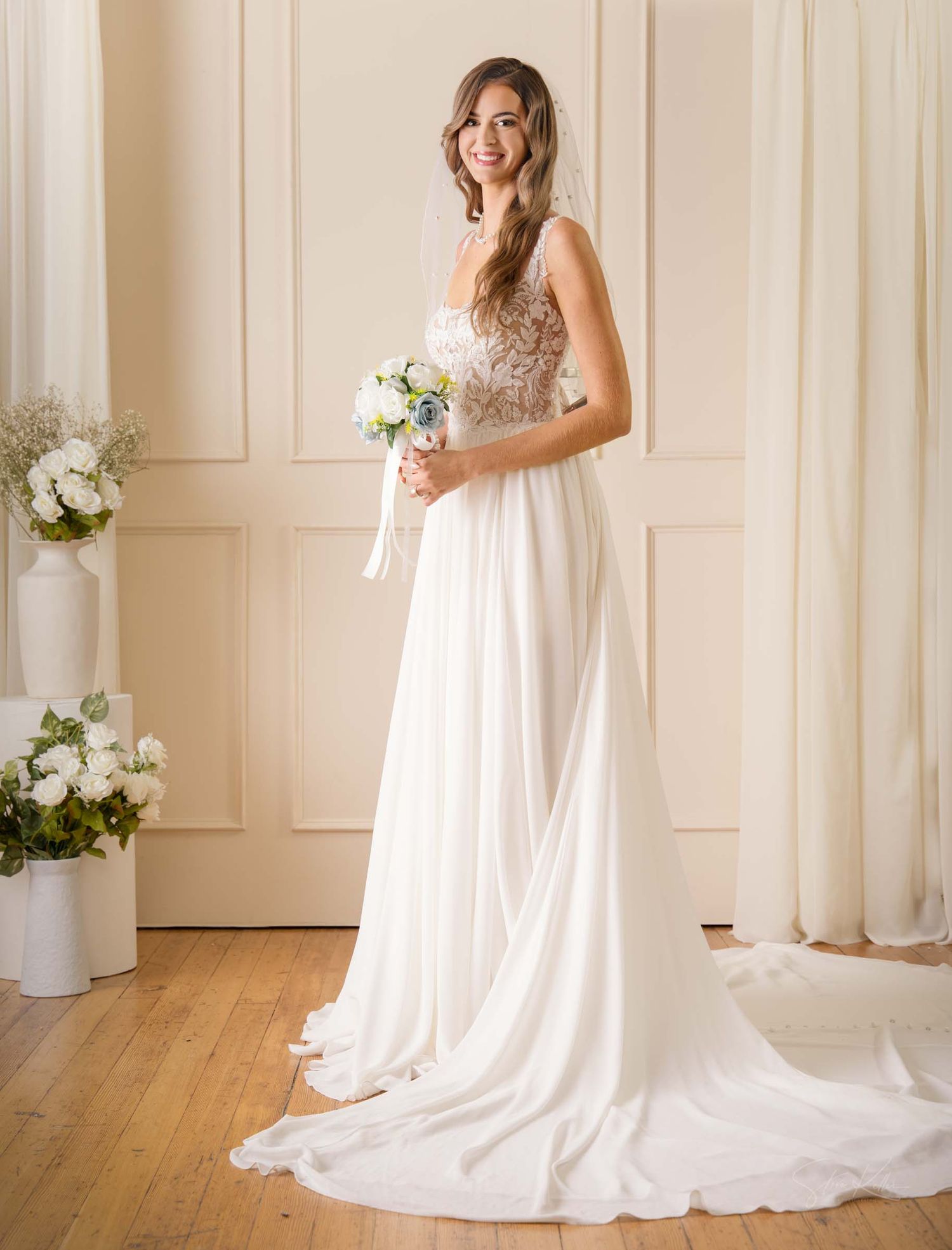 Elegant wedding gown with lace bodice and flowing train displayed in light-filled room with floral decorations.