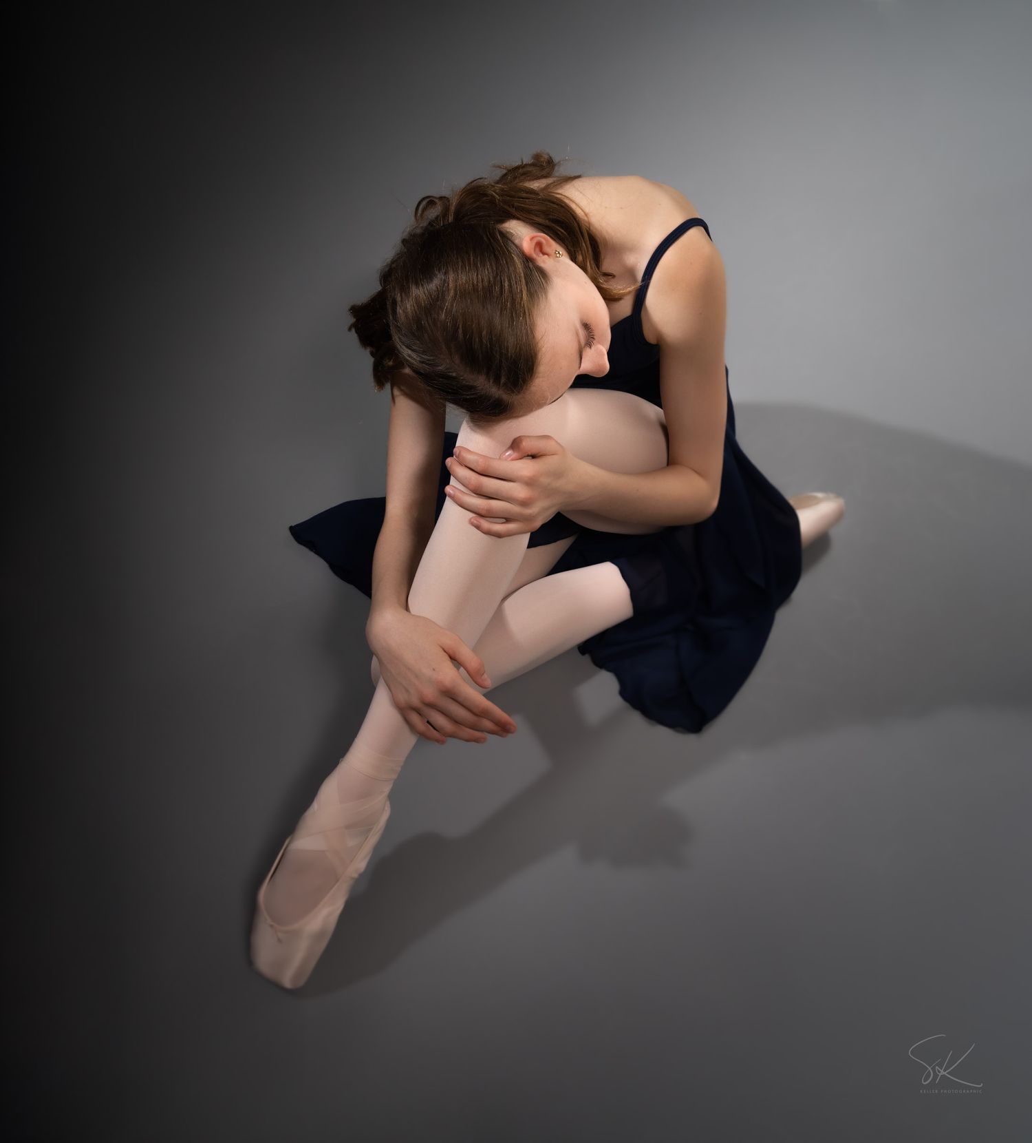 A young ballerina in a navy leotard and pink tights sits gracefully curled on a gray studio floor.