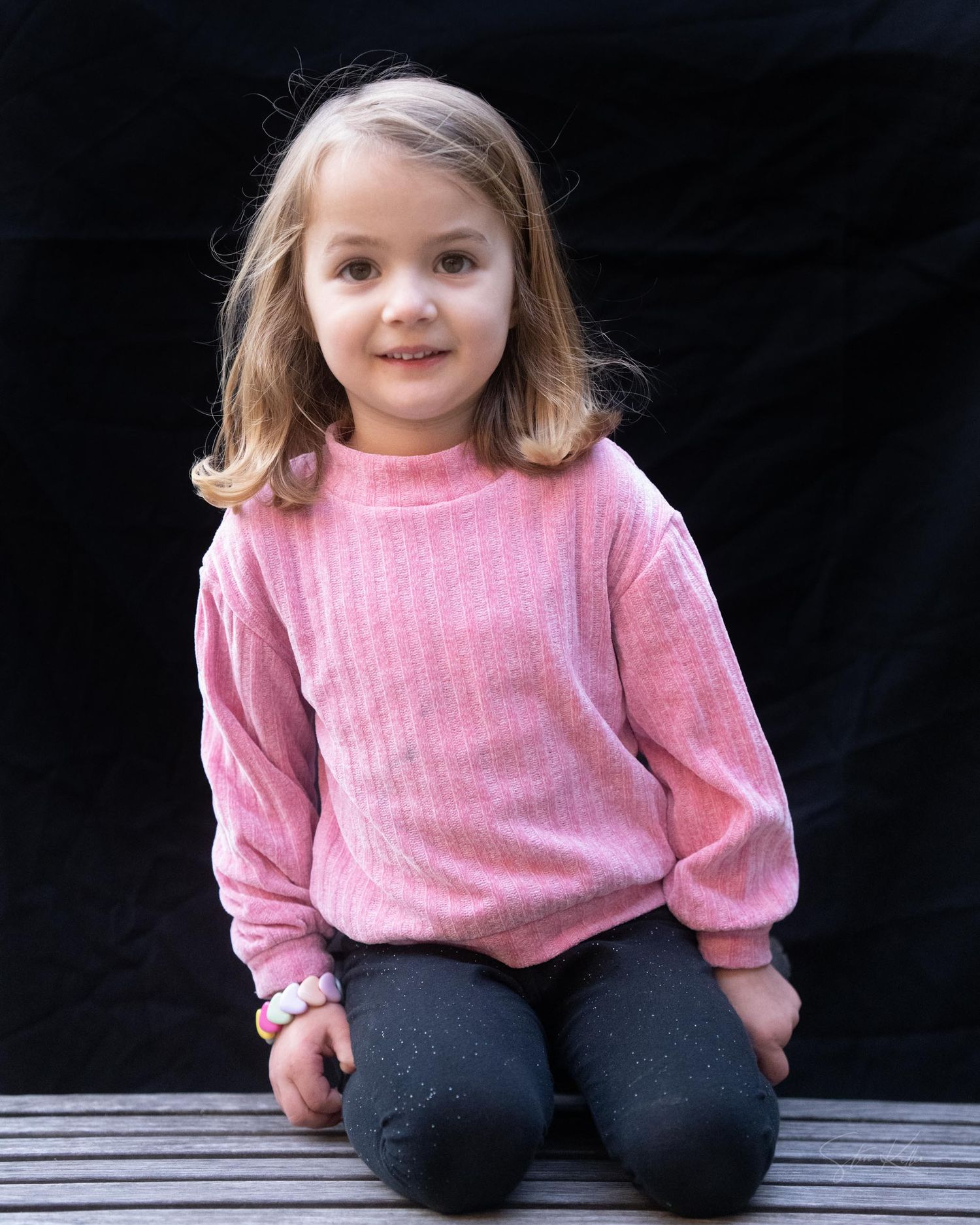 A young child in a pink sweater and dark pants sits casually on a wooden bench against a black background.