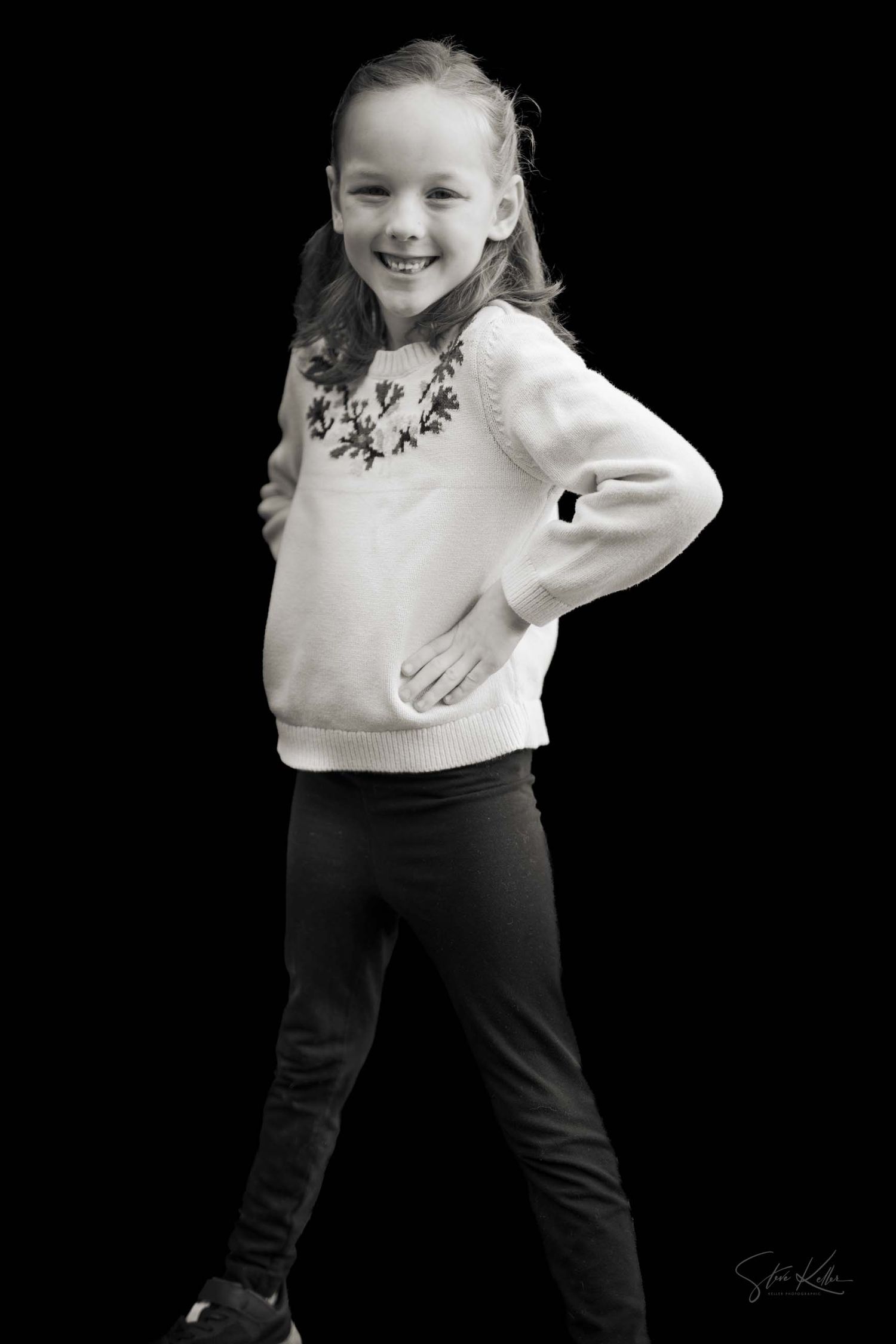 A child in a white sweater and black pants poses playfully against a dark background.