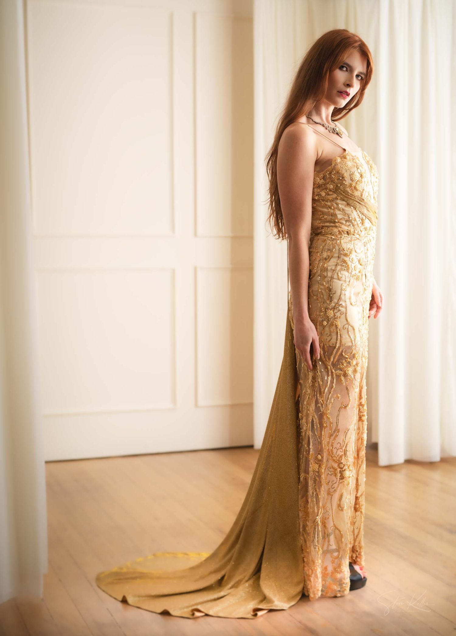 Elegant gold sequined evening gown with flowing train photographed in bright white room with hardwood floors.