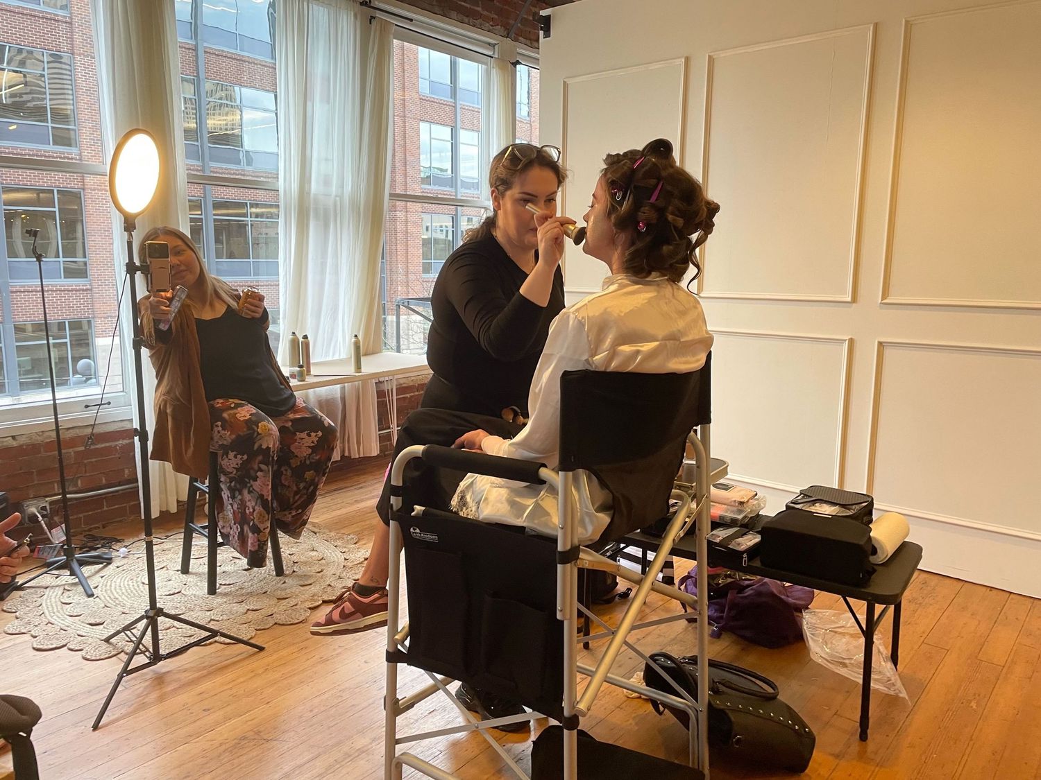 Professional makeup artist applying makeup to a client in a bright studio with ring light and brick backdrop.