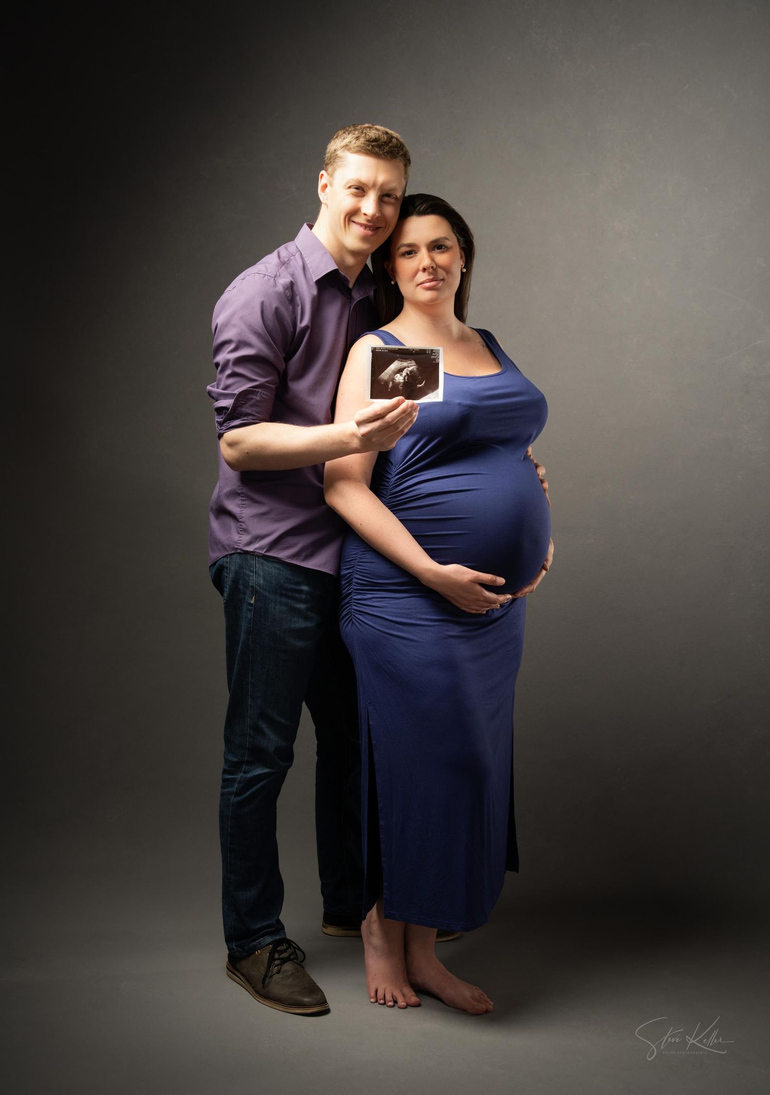 A couple poses with a sonogram photo during a maternity photoshoot, with the expectant mother wearing a navy blue dress.