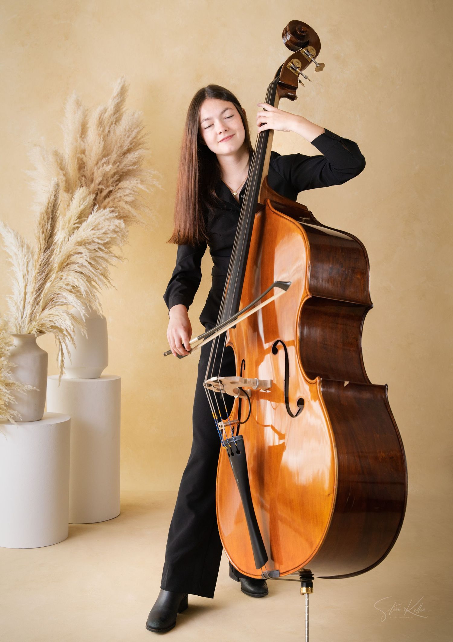 A musician in black attire plays a wooden double bass next to decorative pampas grass against a beige backdrop.