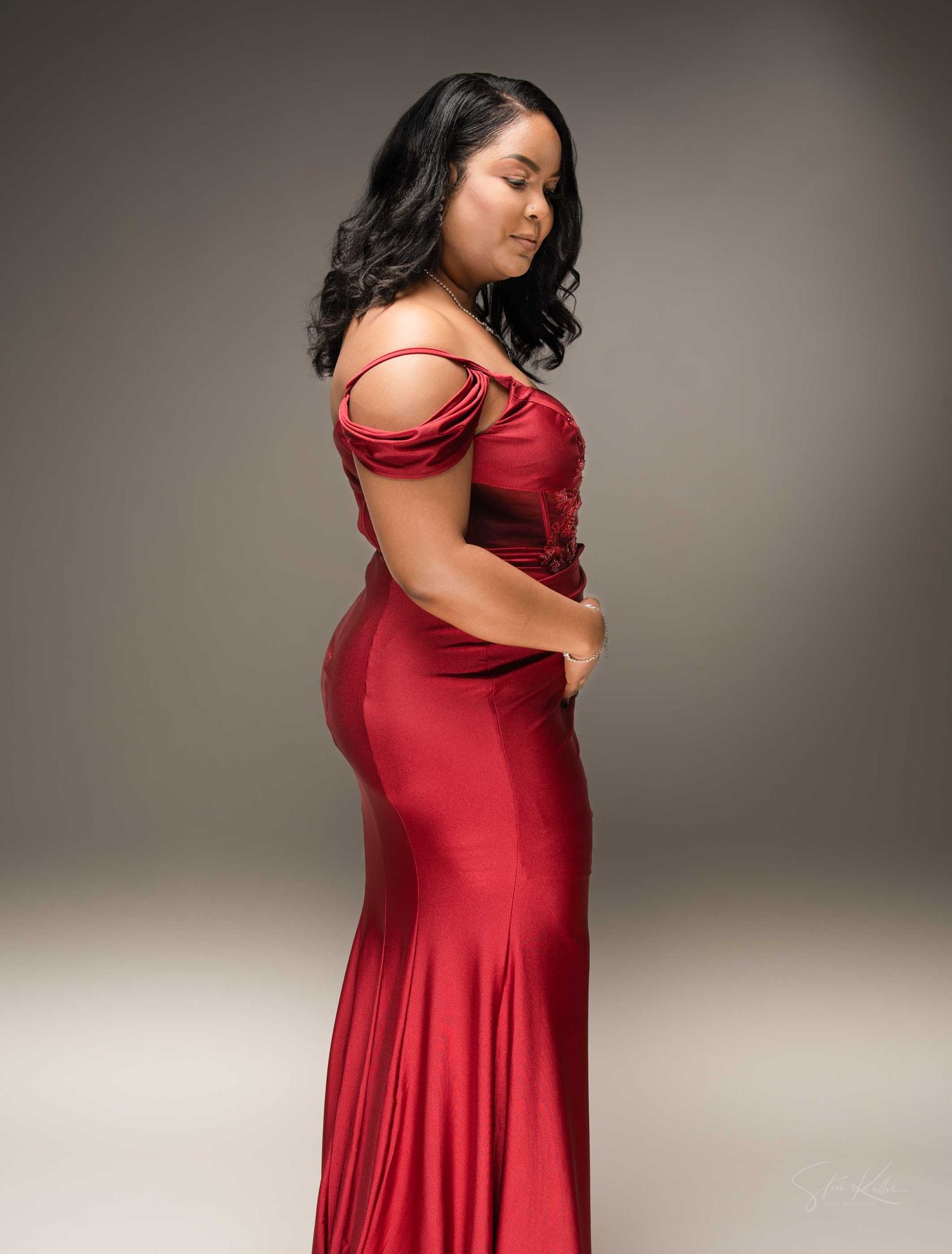 Woman in elegant red satin off-shoulder mermaid gown posing gracefully in a professional studio setting.