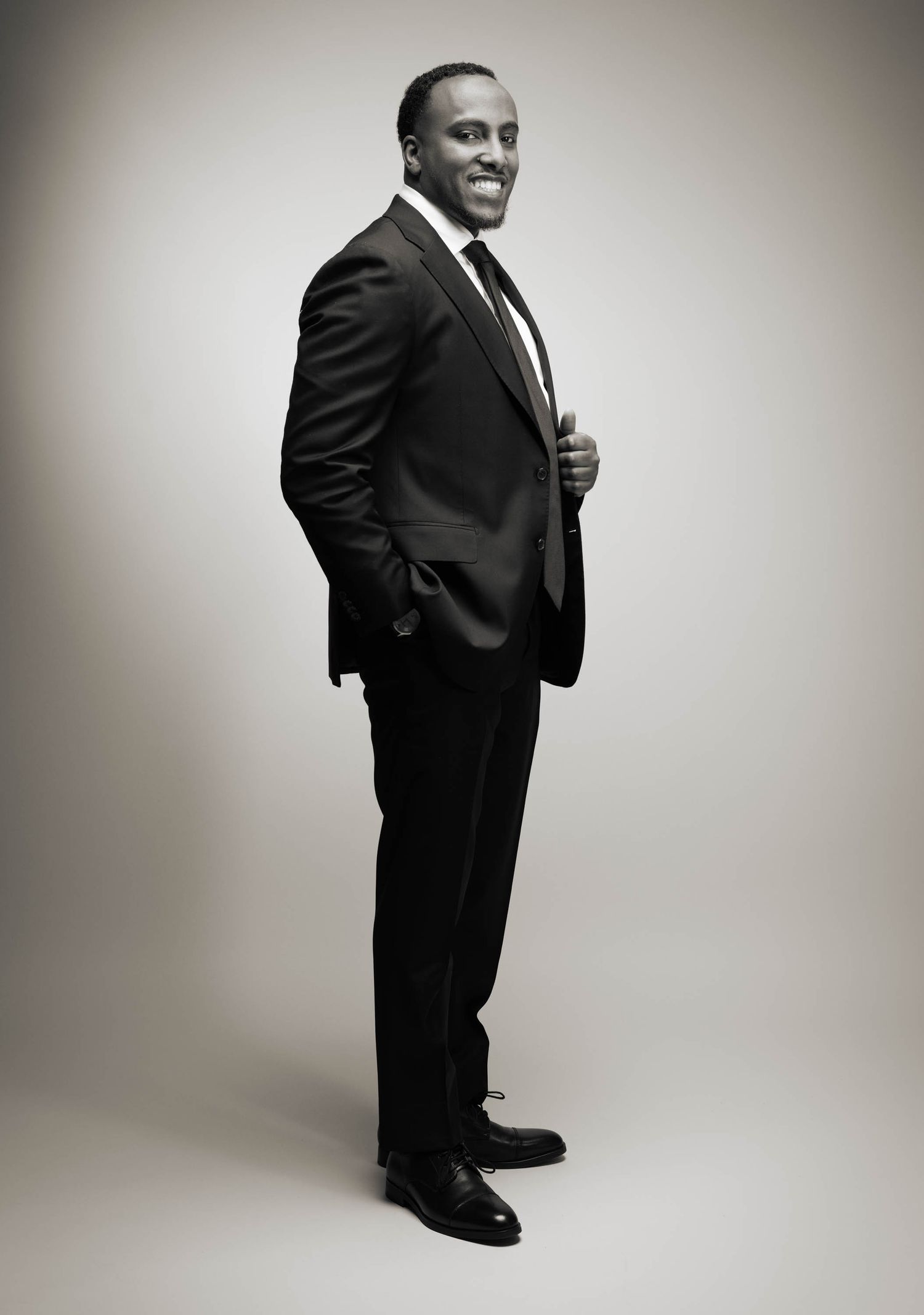 Professional black and white studio portrait of a figure in a sleek dark suit and tie standing confidently against a gradient background.