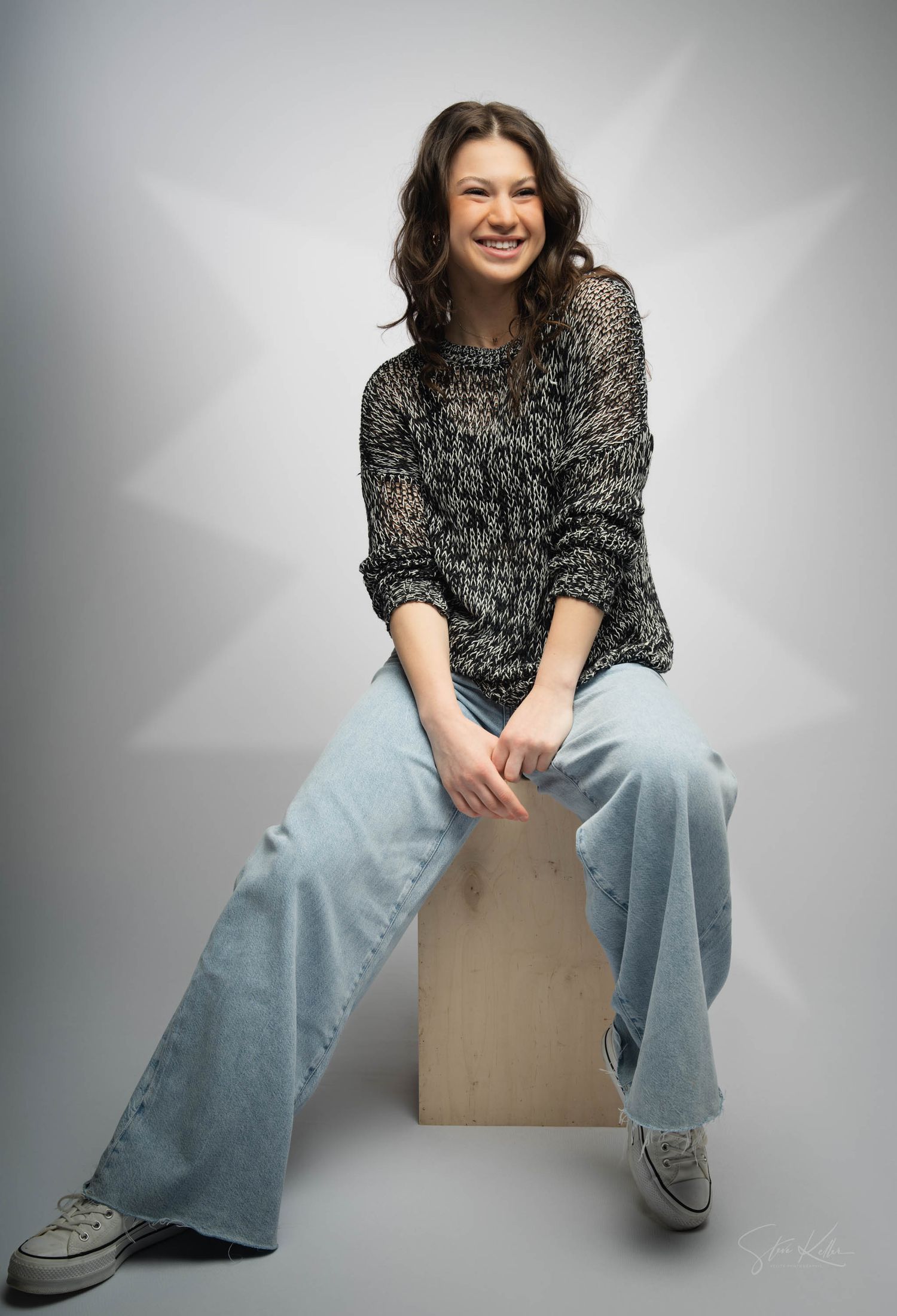 fashion model in a casual knit sweater and wide-leg jeans sits on a cube against a light gray studio background.