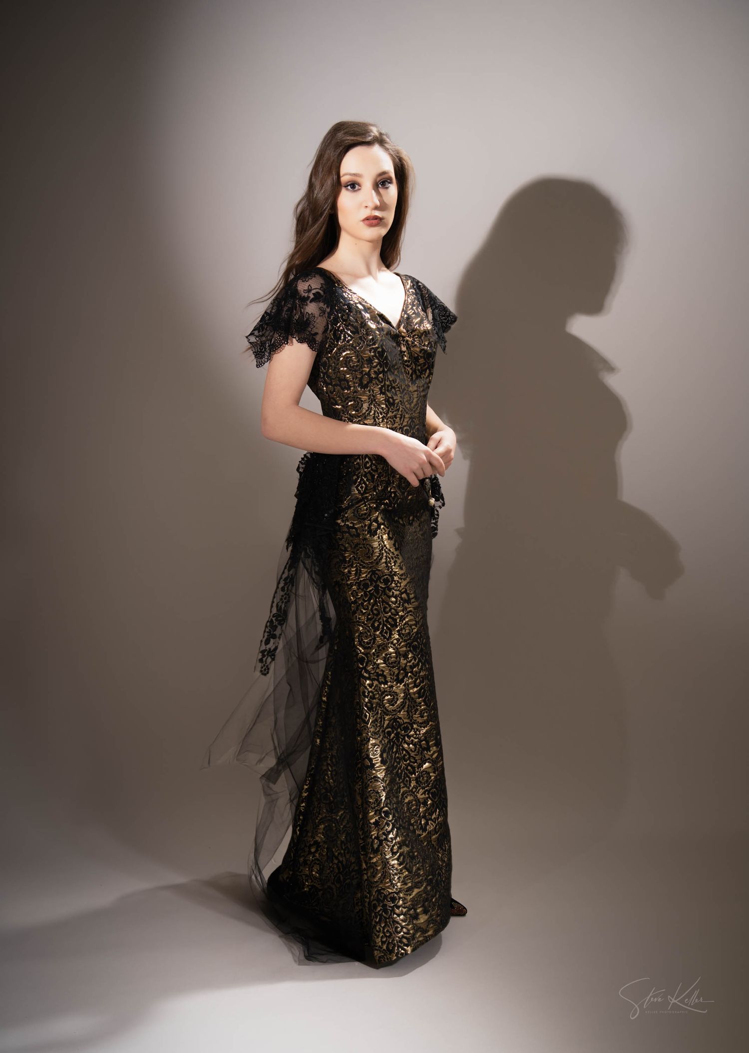 A model in a black and gold lace evening gown with short sleeves poses dramatically against a neutral backdrop.