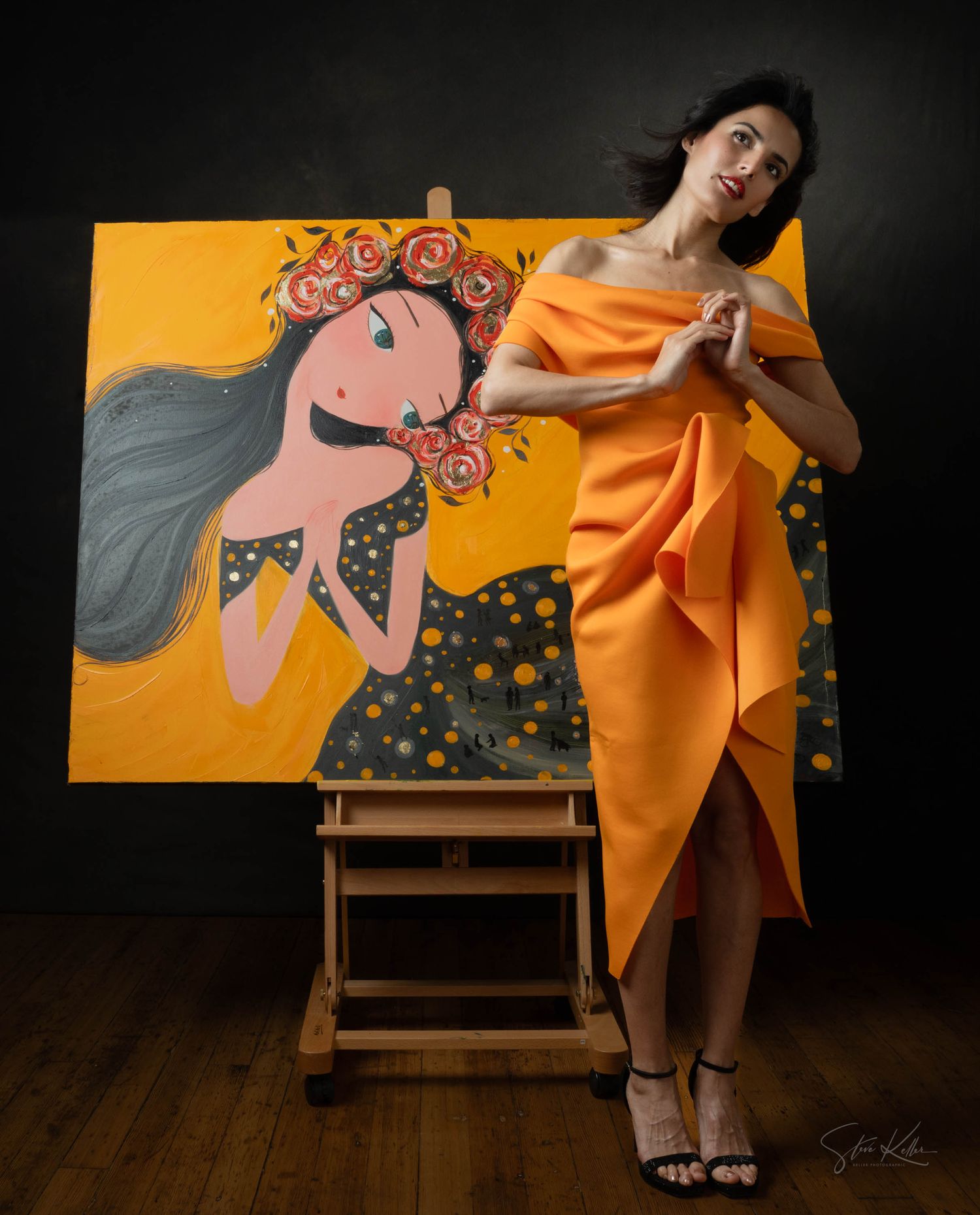 Artist poses dramatically next to a vibrant painting of a whimsical figure with flowers on an orange background on an easel.