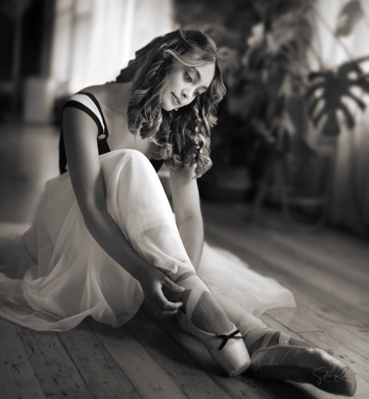 A graceful dancer in a white dress performs an elegant pose on a wooden floor in black and white.