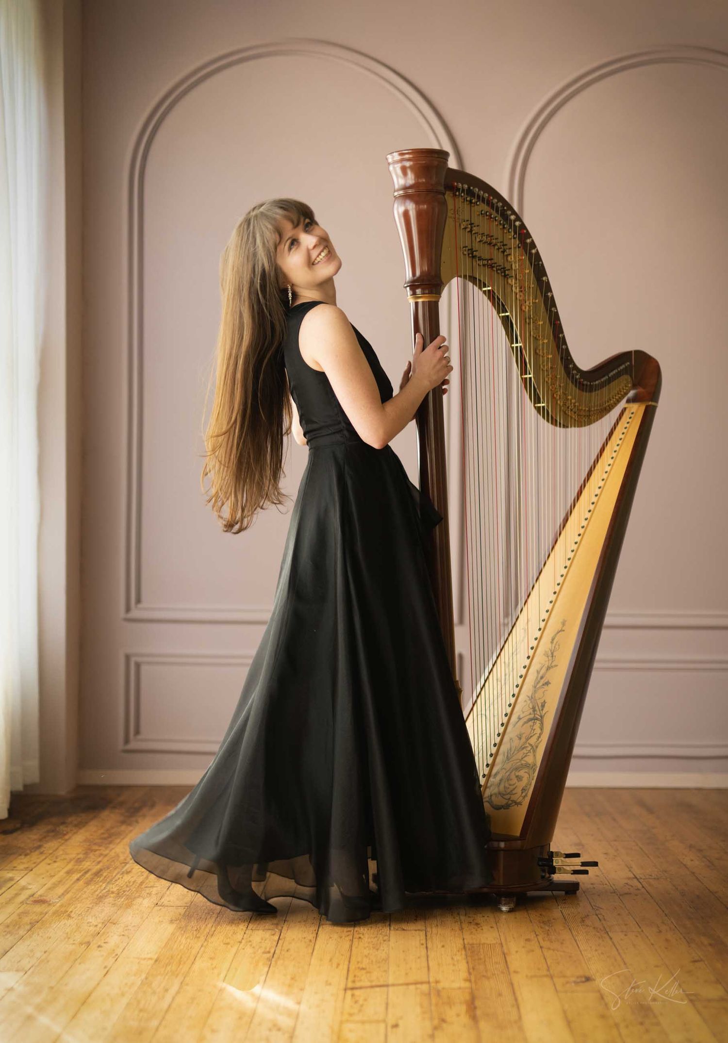 Smiling female harpist in black gown posing elegantly with a large concert harp on a wooden floor.