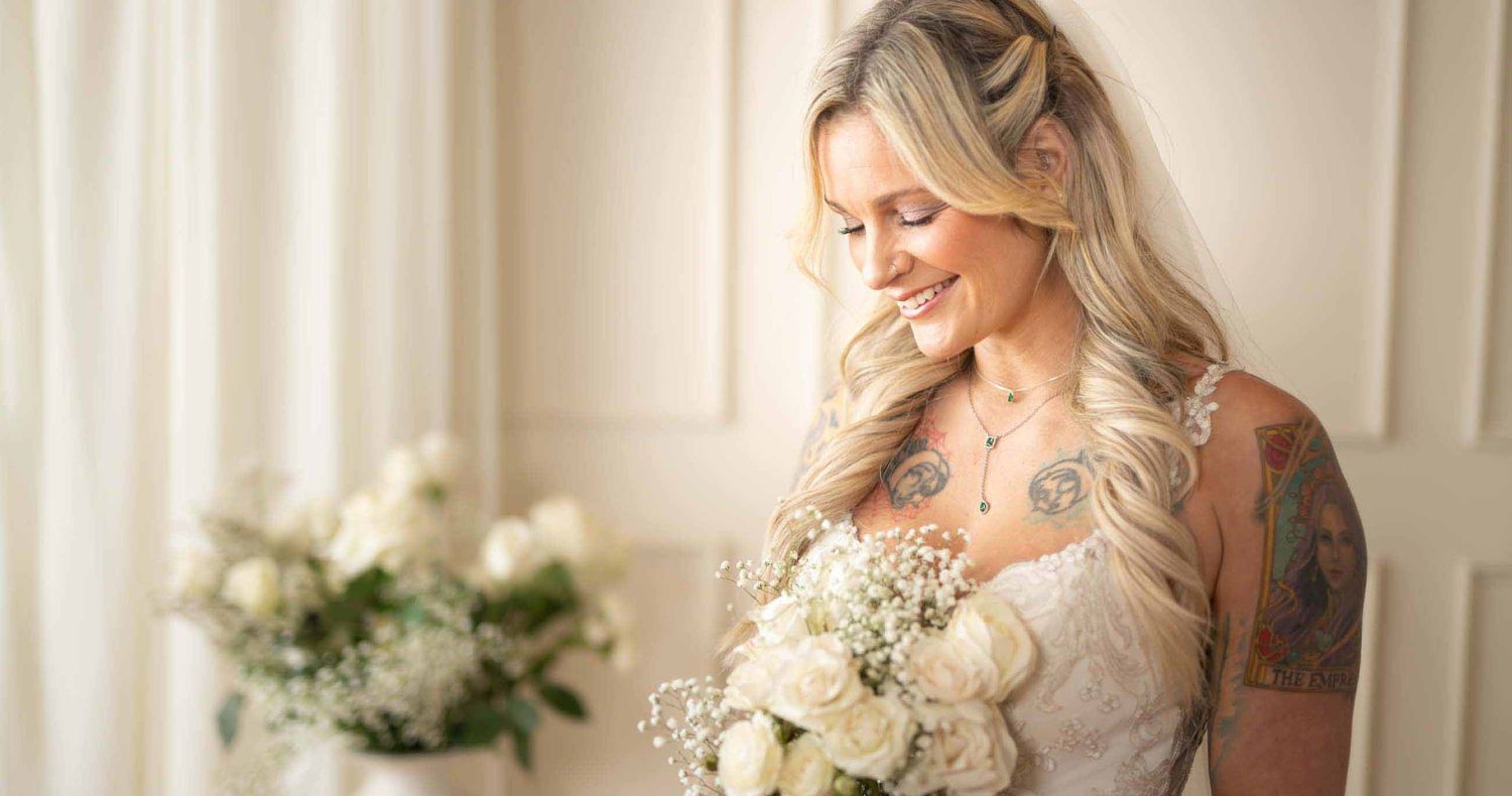 Smiling tattooed bride with long wavy blonde hair holds a white floral bouquet in a lace wedding dress beside soft curtains.