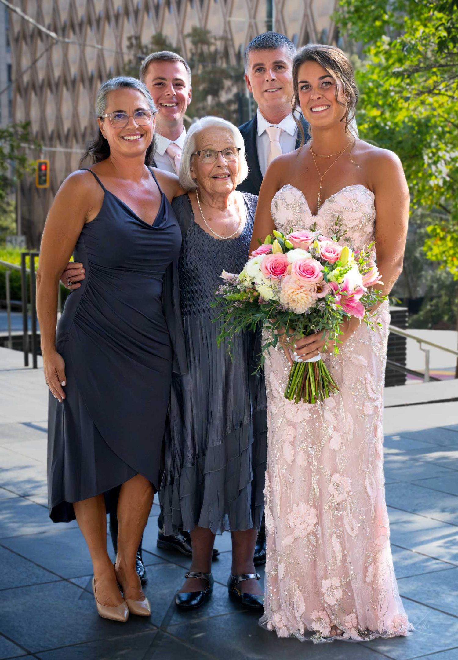 A bride in a lace gown holds pink roses bouquet posing with family including an elderly woman at a wedding.