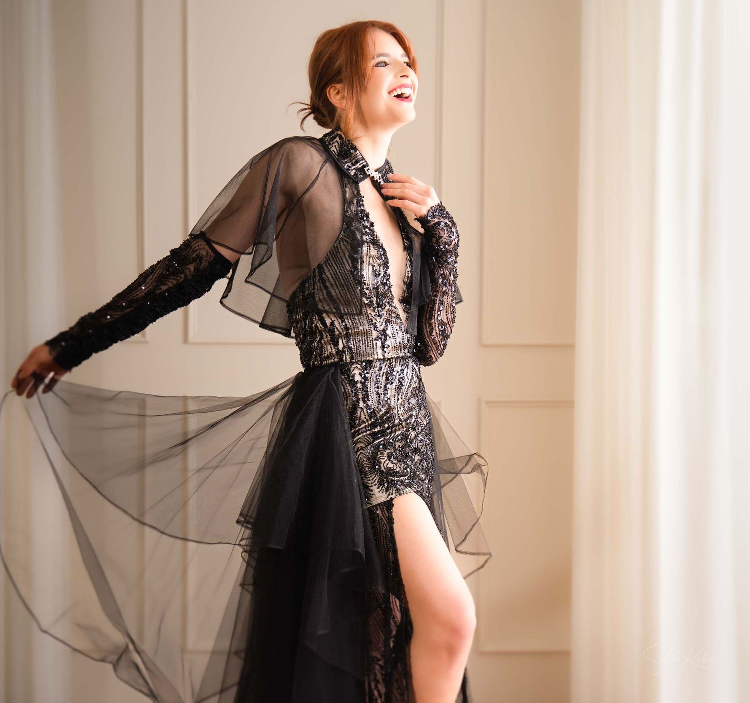 Woman in black lace dress with flowing sheer sleeves poses gracefully against neutral wall.