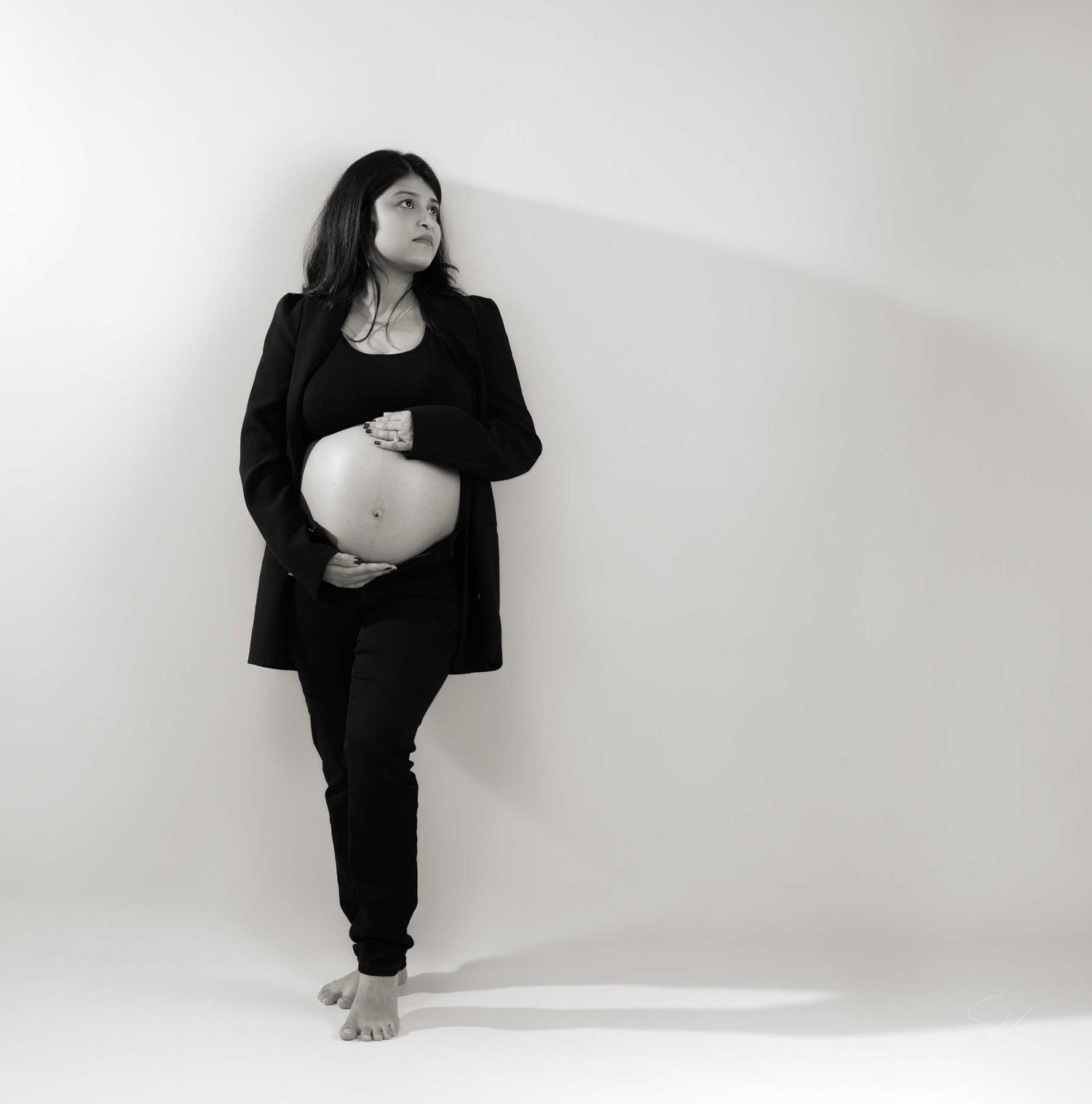 Black and white portrait of a pregnant woman in black outfit, barefoot, leaning against white wall cradling her belly.
