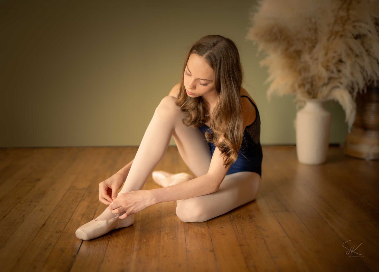 A young ballerina in a blue leotard ties her pointe shoes while sitting gracefully on a wooden floor.