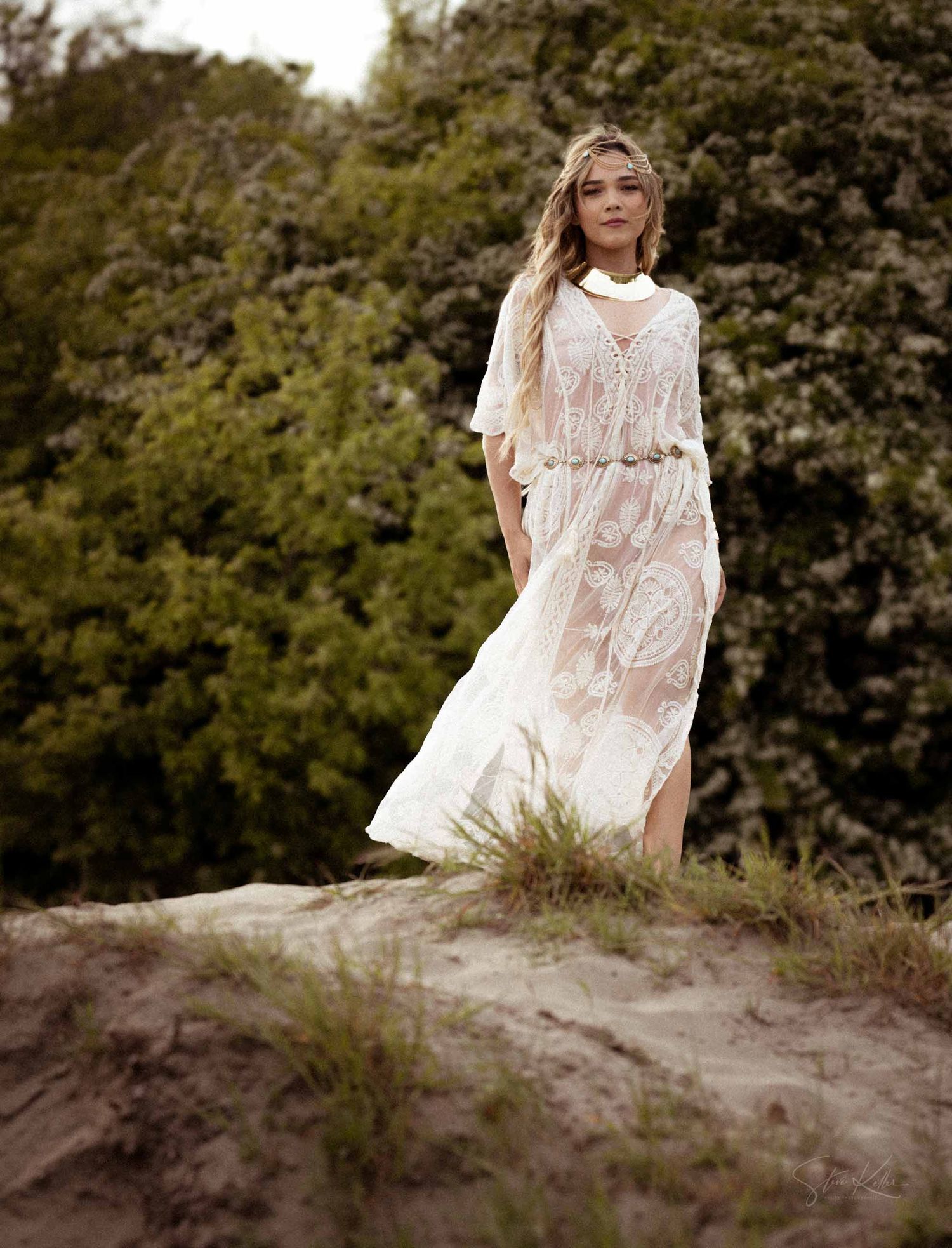 A figure in a flowing white dress poses gracefully on a grassy hillside against a natural wooded backdrop.