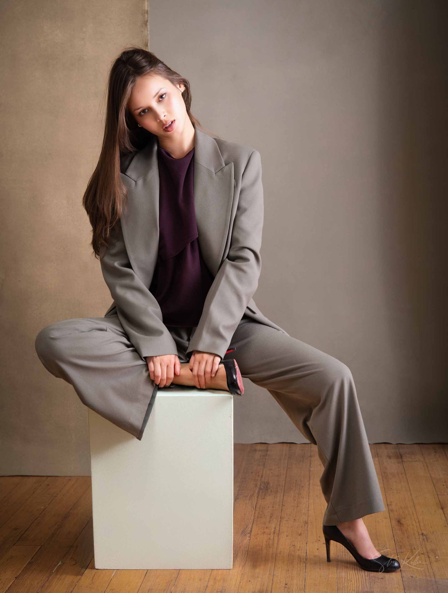 Professional portrait of someone in a gray business suit and purple top seated casually on a white cube against a neutral wall.