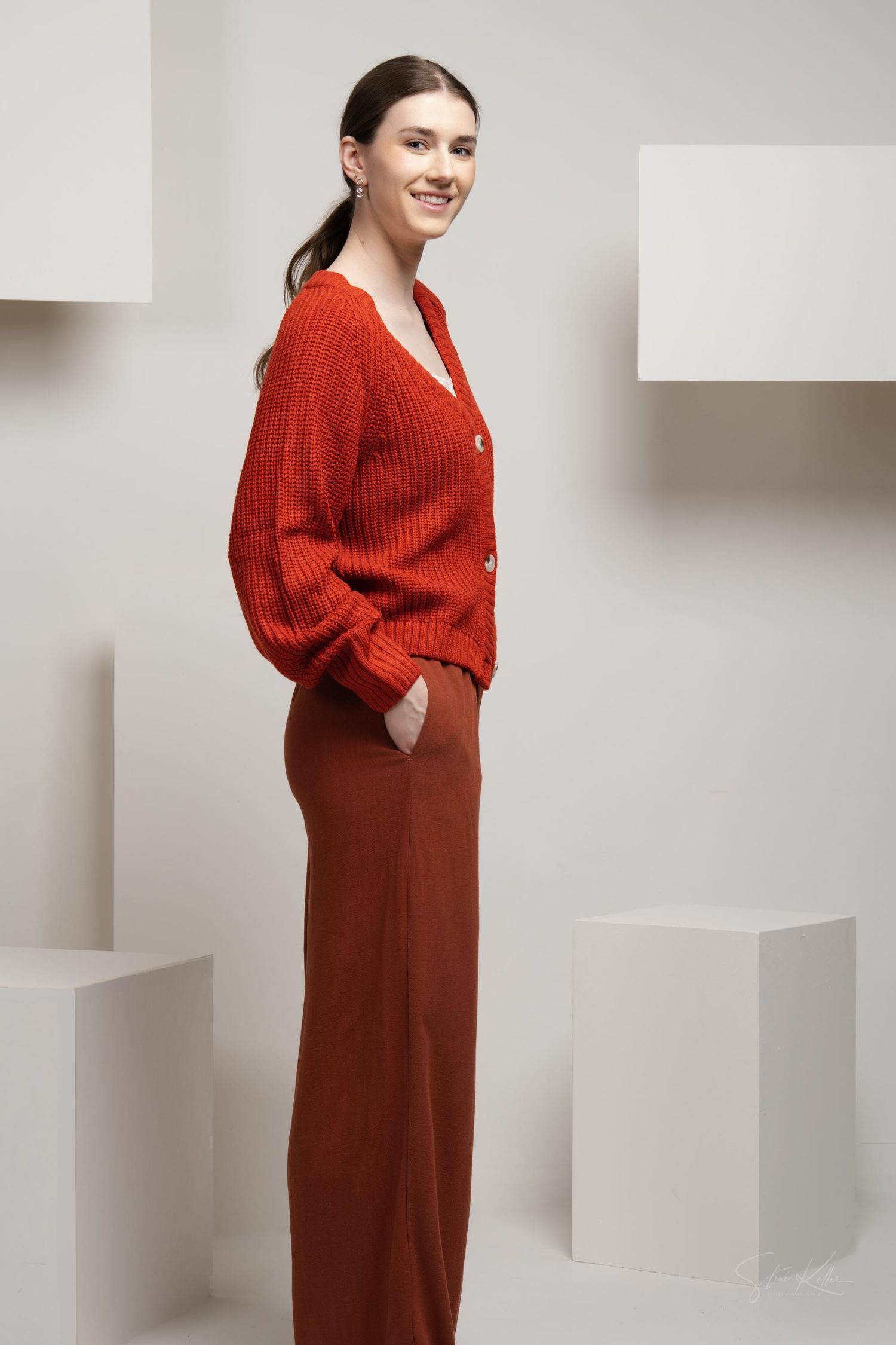 Model wearing a cozy red knit cardigan and matching wide-leg pants poses elegantly against white geometric background.