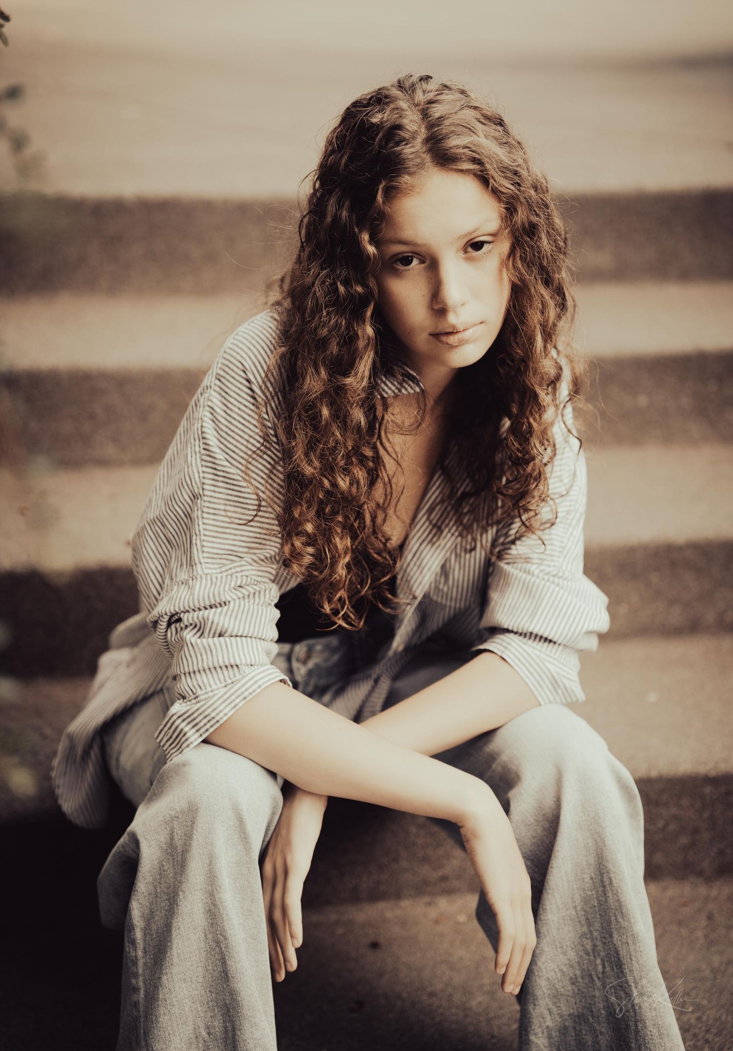 A person with long curly brown hair sits on stone steps wearing an oversized sweater in a vintage-toned photograph.