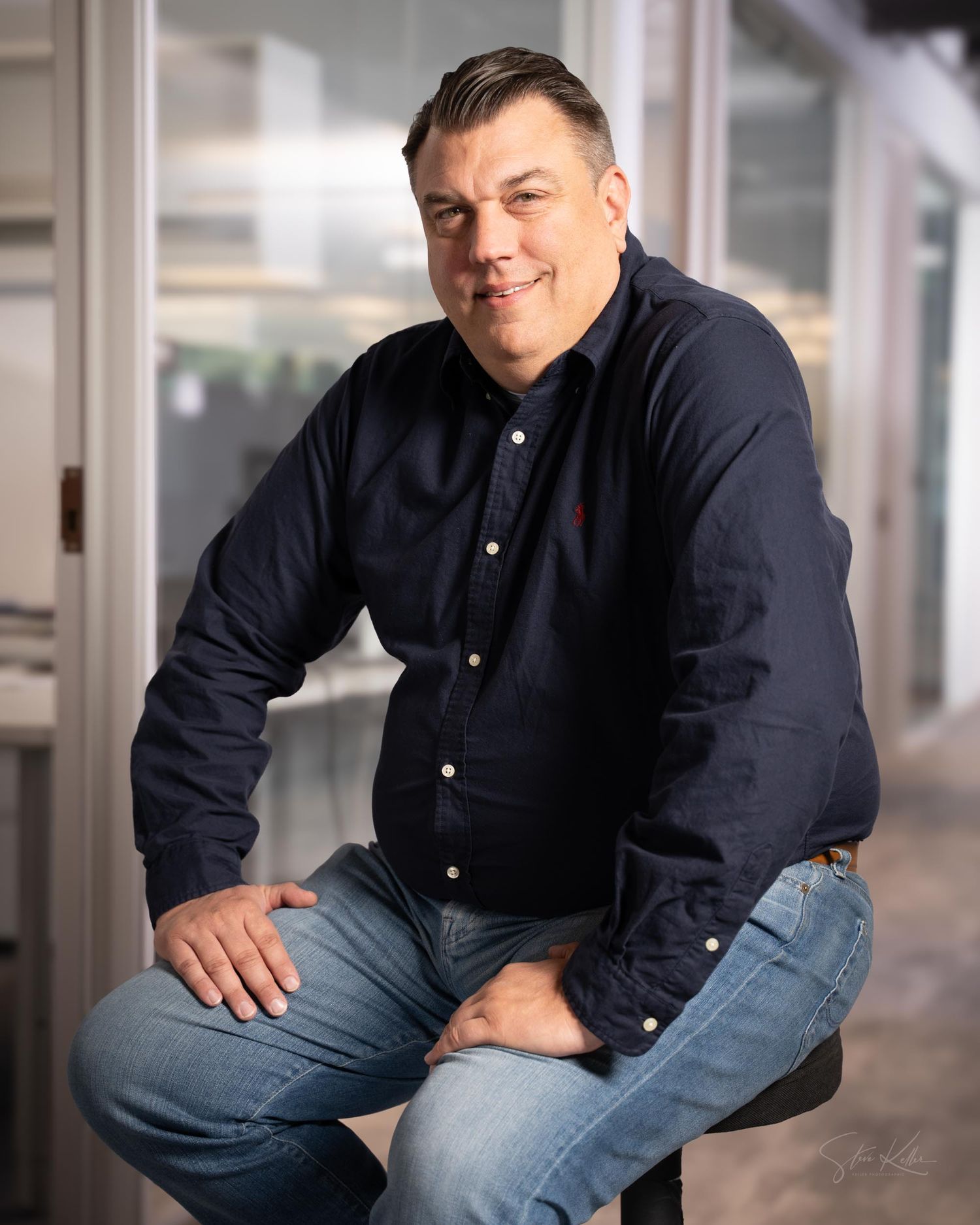 A person wearing a navy button-down shirt and jeans sits casually in a studio setting with a white background.